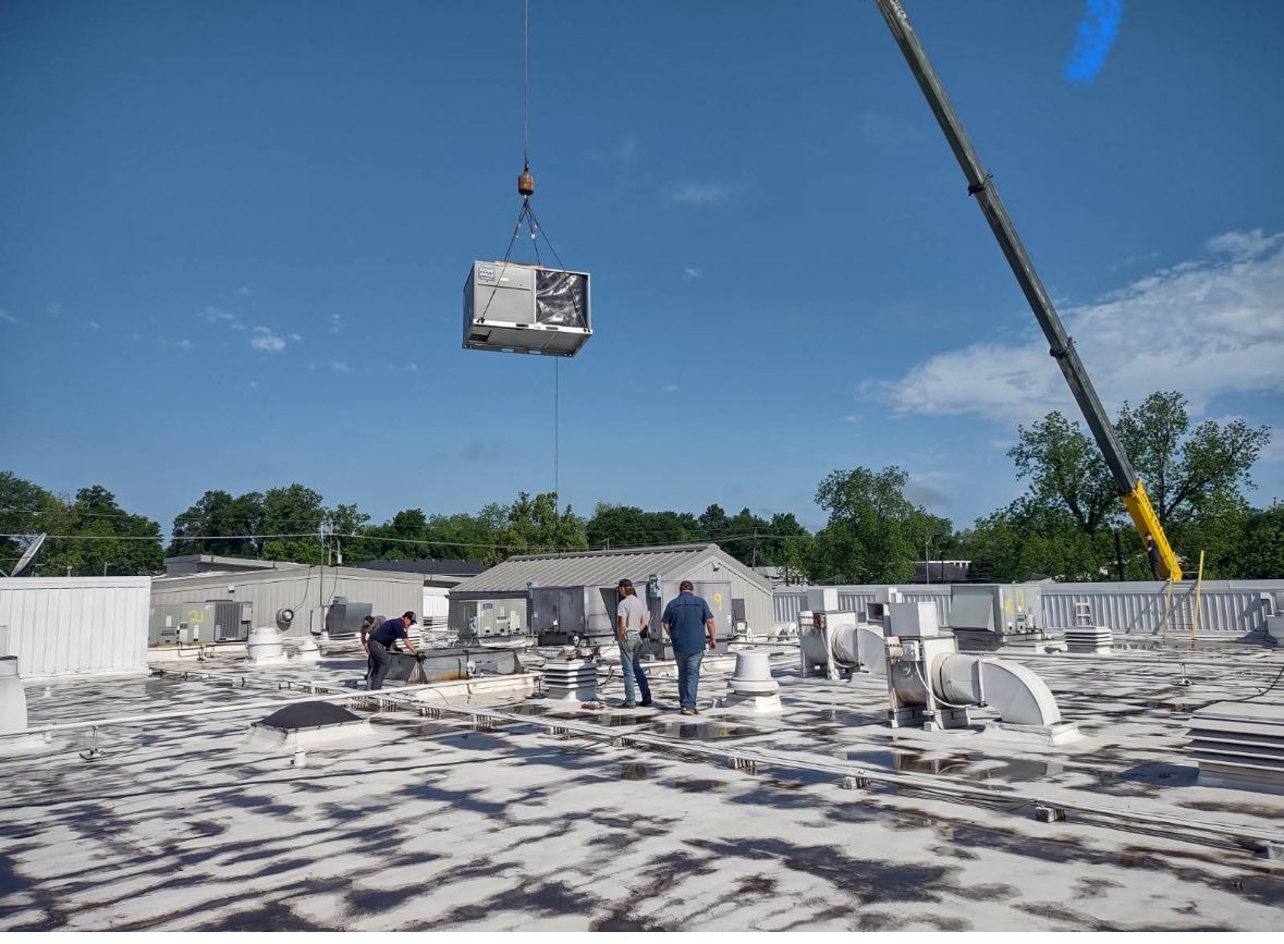 A crane lifting an HVAC unit onto a flat commercial roof, where workers are present.