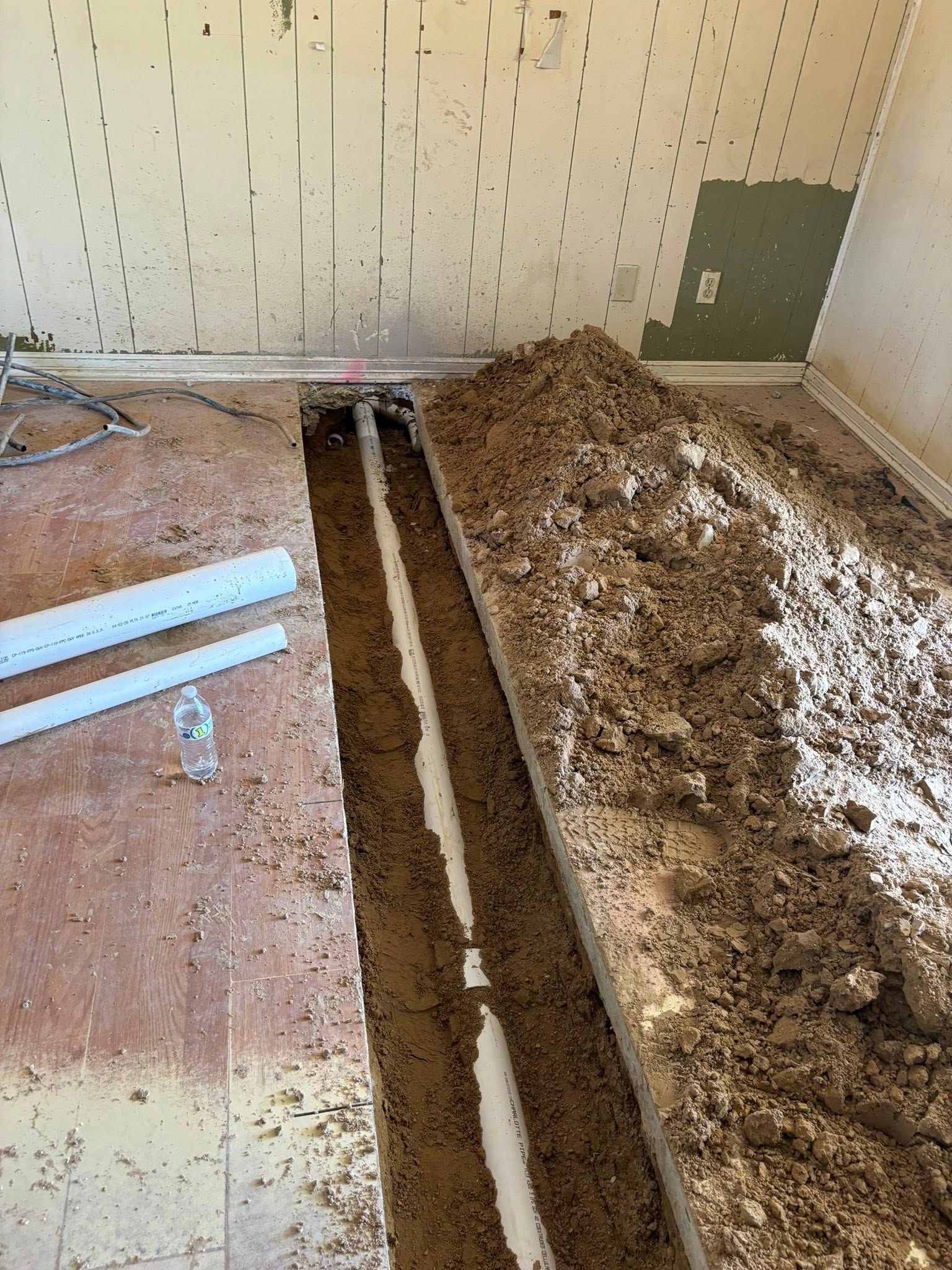 Trench dug in floor with white pipe visible. Dirt piled next to it. Wall damaged.