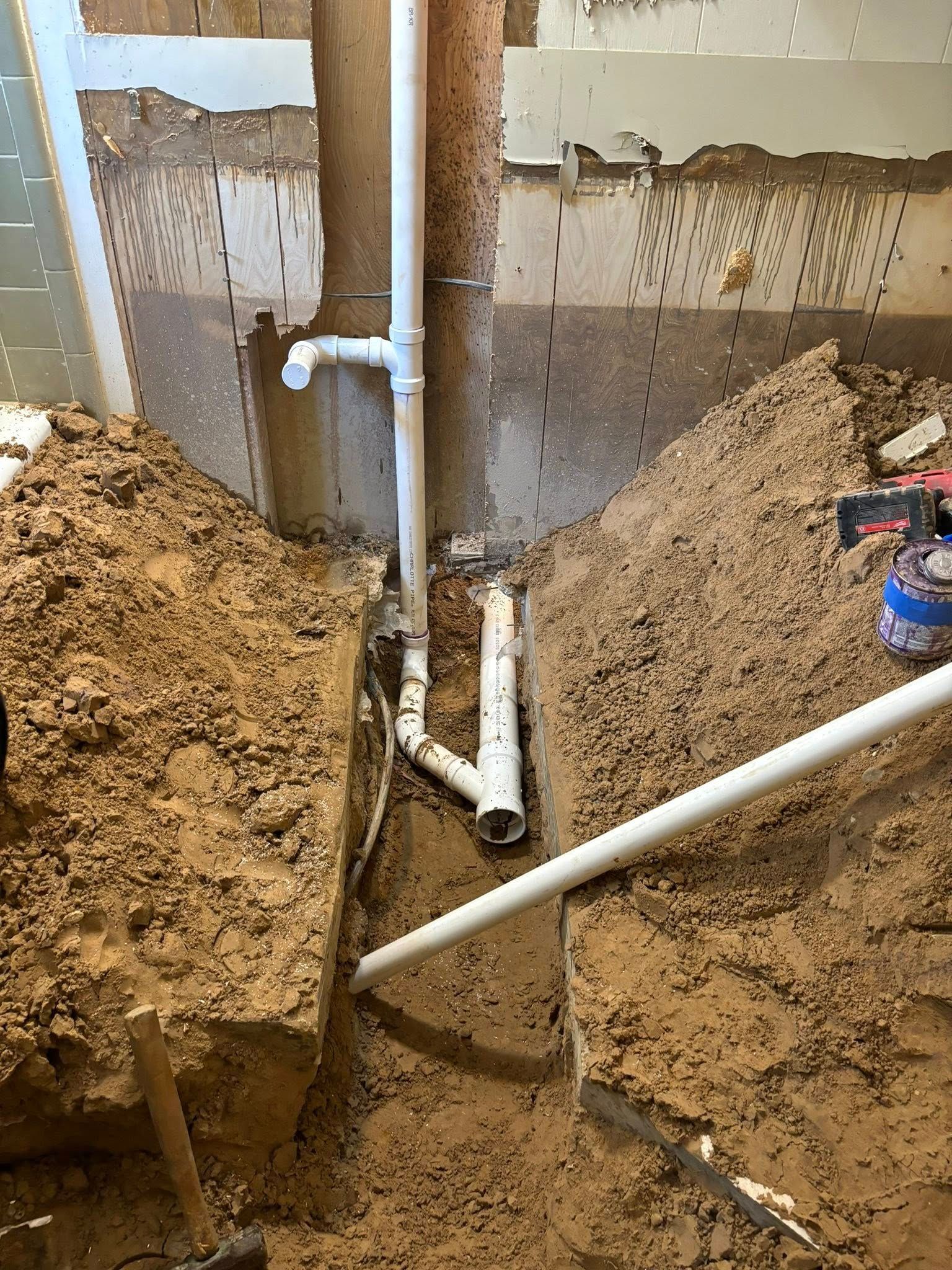 Excavated plumbing pipes in dirt trench near a concrete wall. White PVC pipes with connections visible.