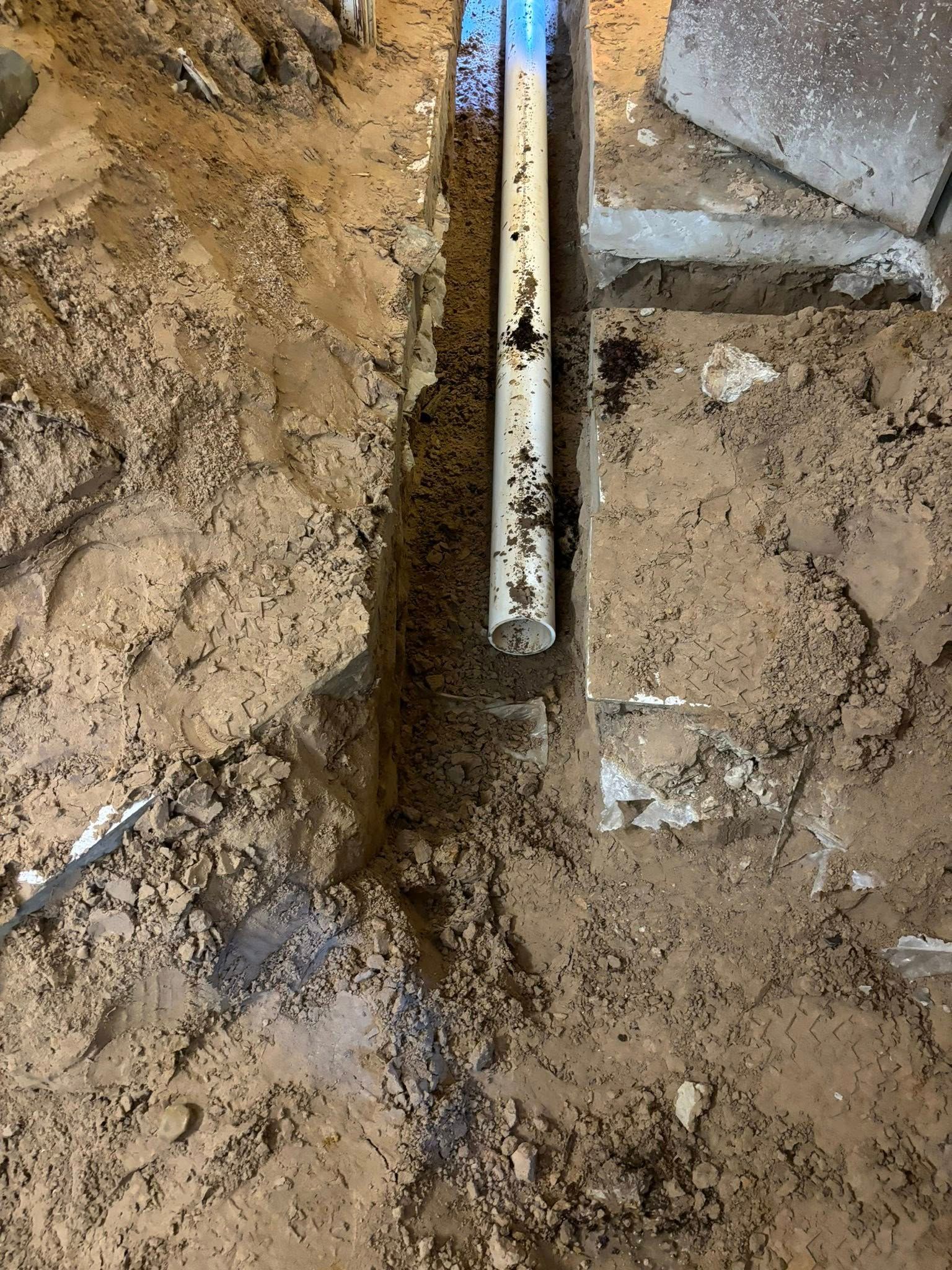 Trench in dirt with white pipe. Partially visible concrete blocks and blue marking tape on pipe.