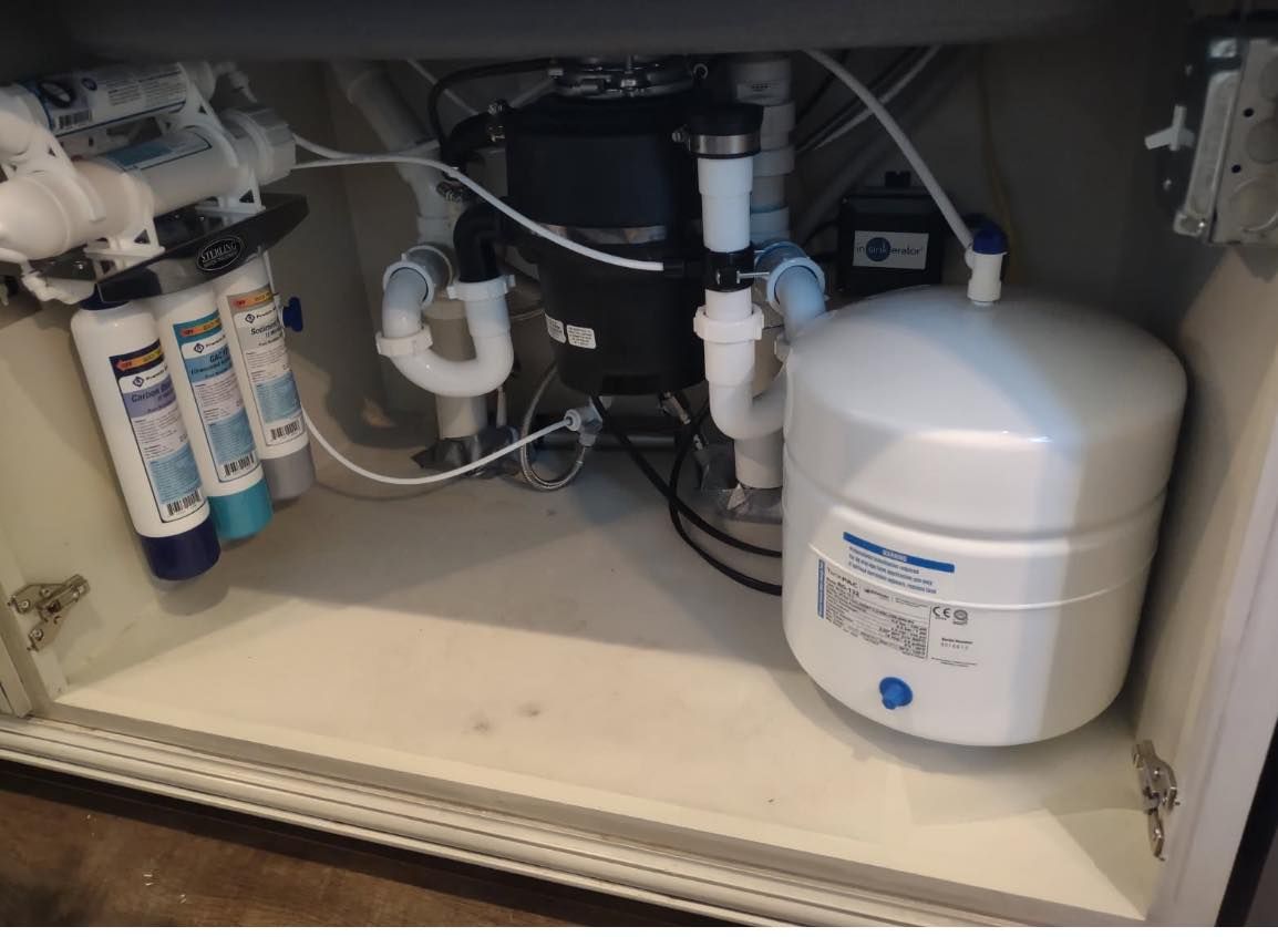 Water filtration system under a sink, with filters, tank, and plumbing visible.