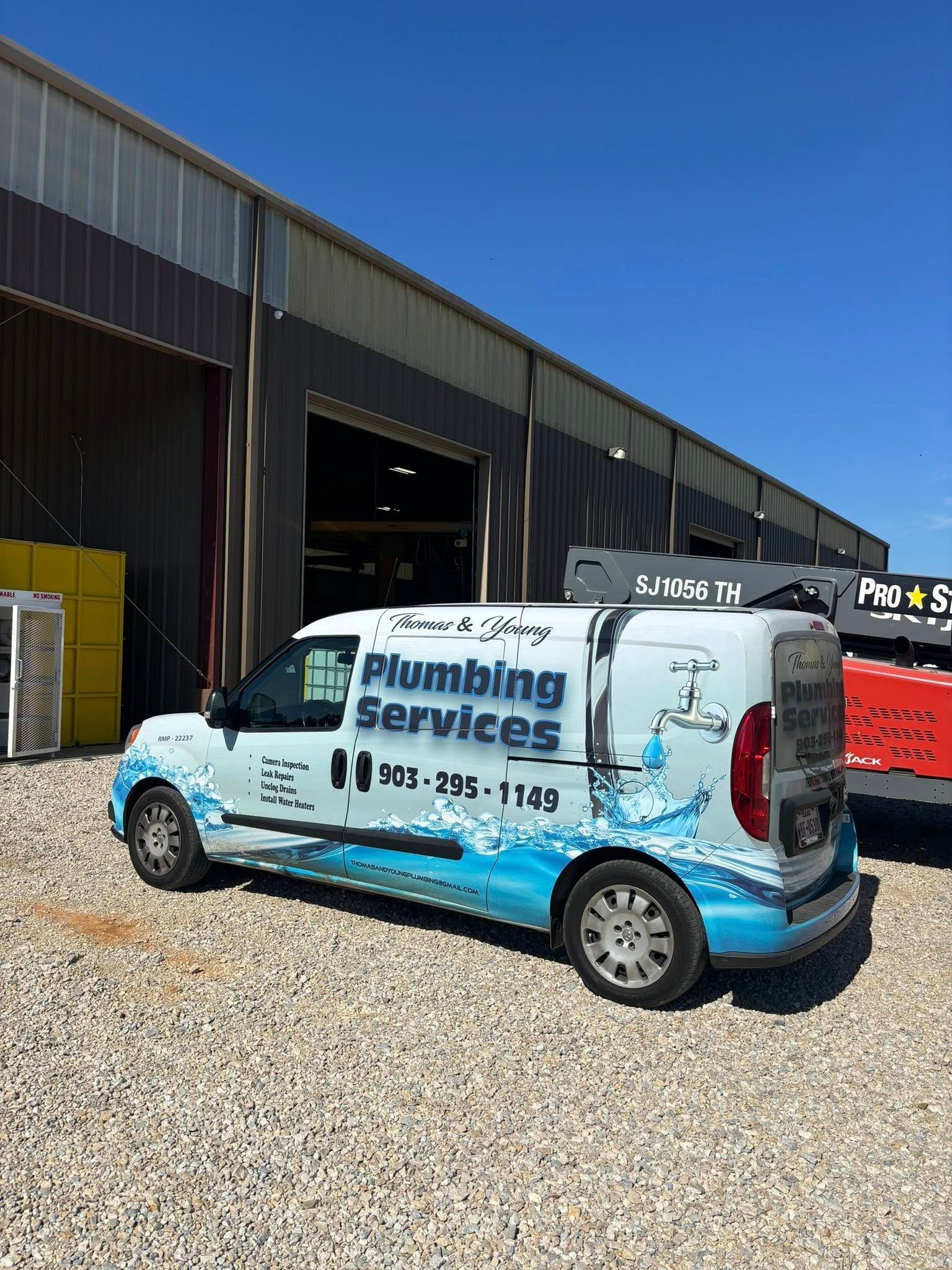 White and blue plumbing service van parked in front of a building on a gravel lot under a blue sky.