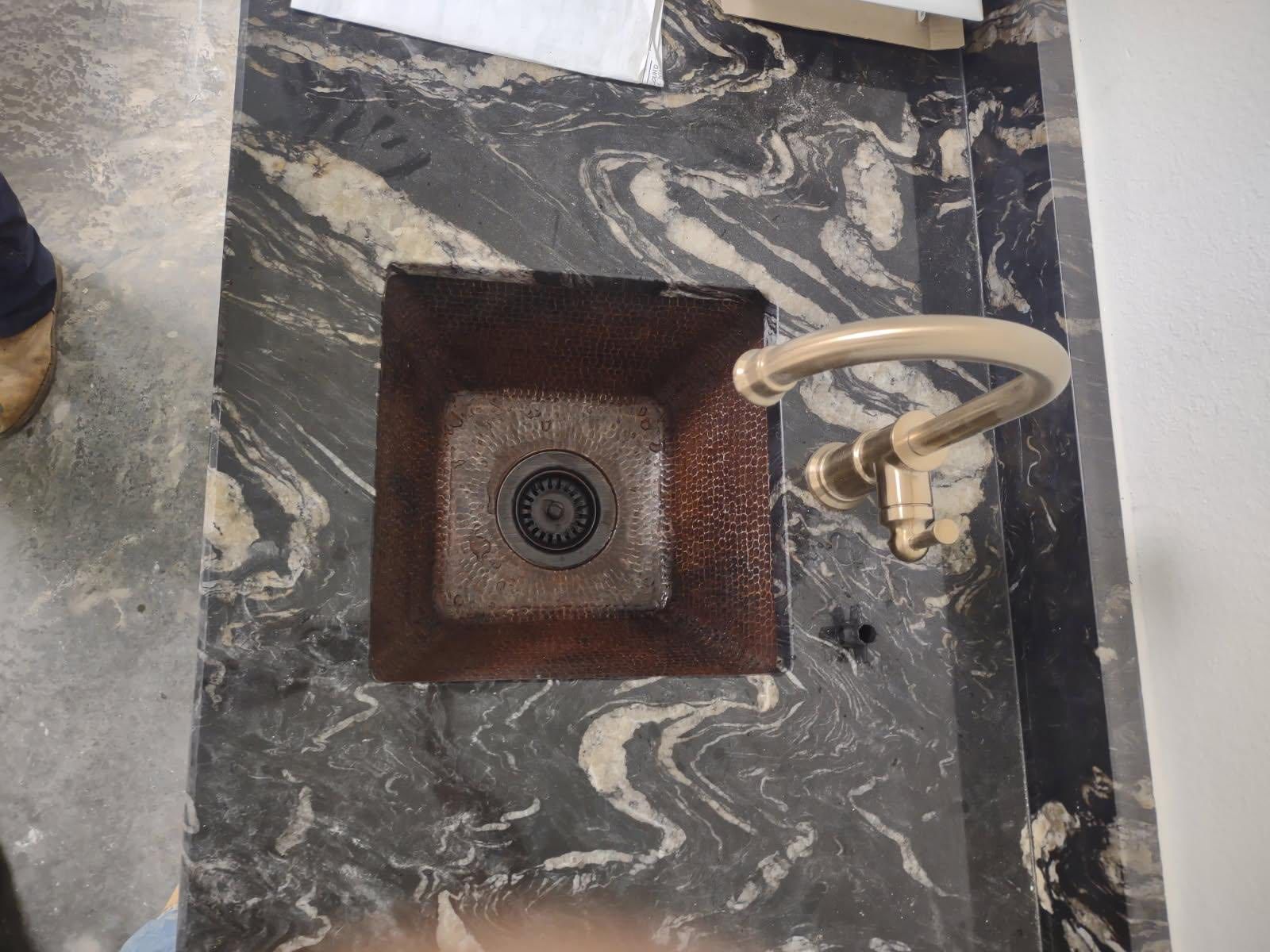 Copper square sink set into dark granite countertop with curved faucet.