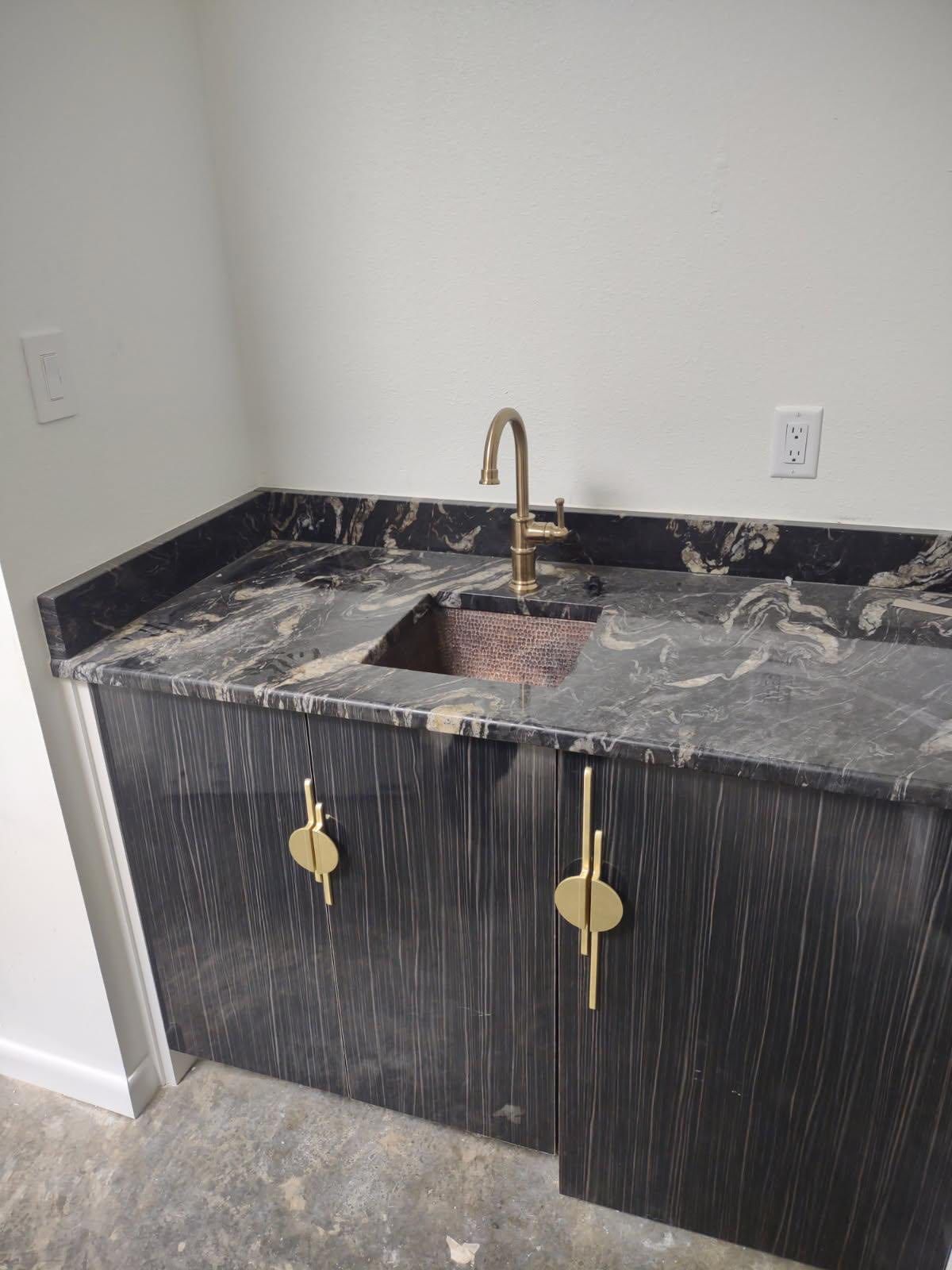 Dark cabinet with gold handles, copper sink, gold faucet, and black granite countertop in a room.