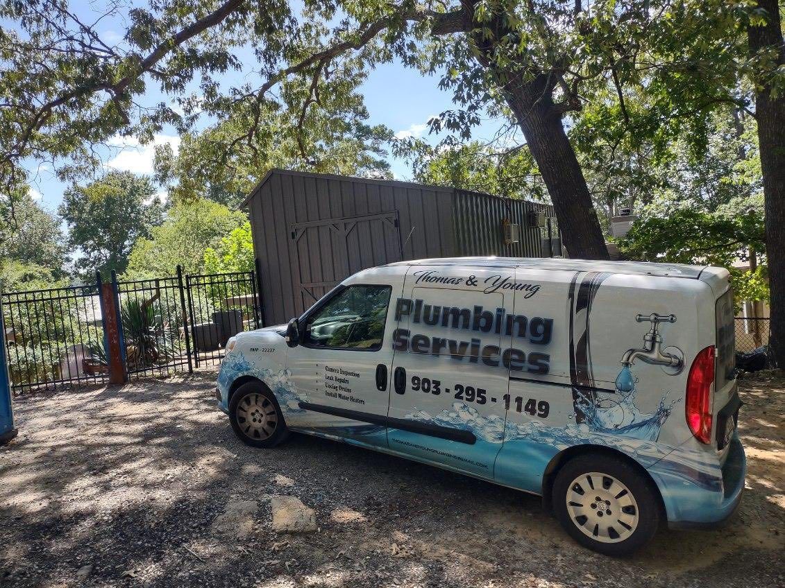 White plumbing service van parked outside a fence, under trees. Van has blue water splash design and phone number.