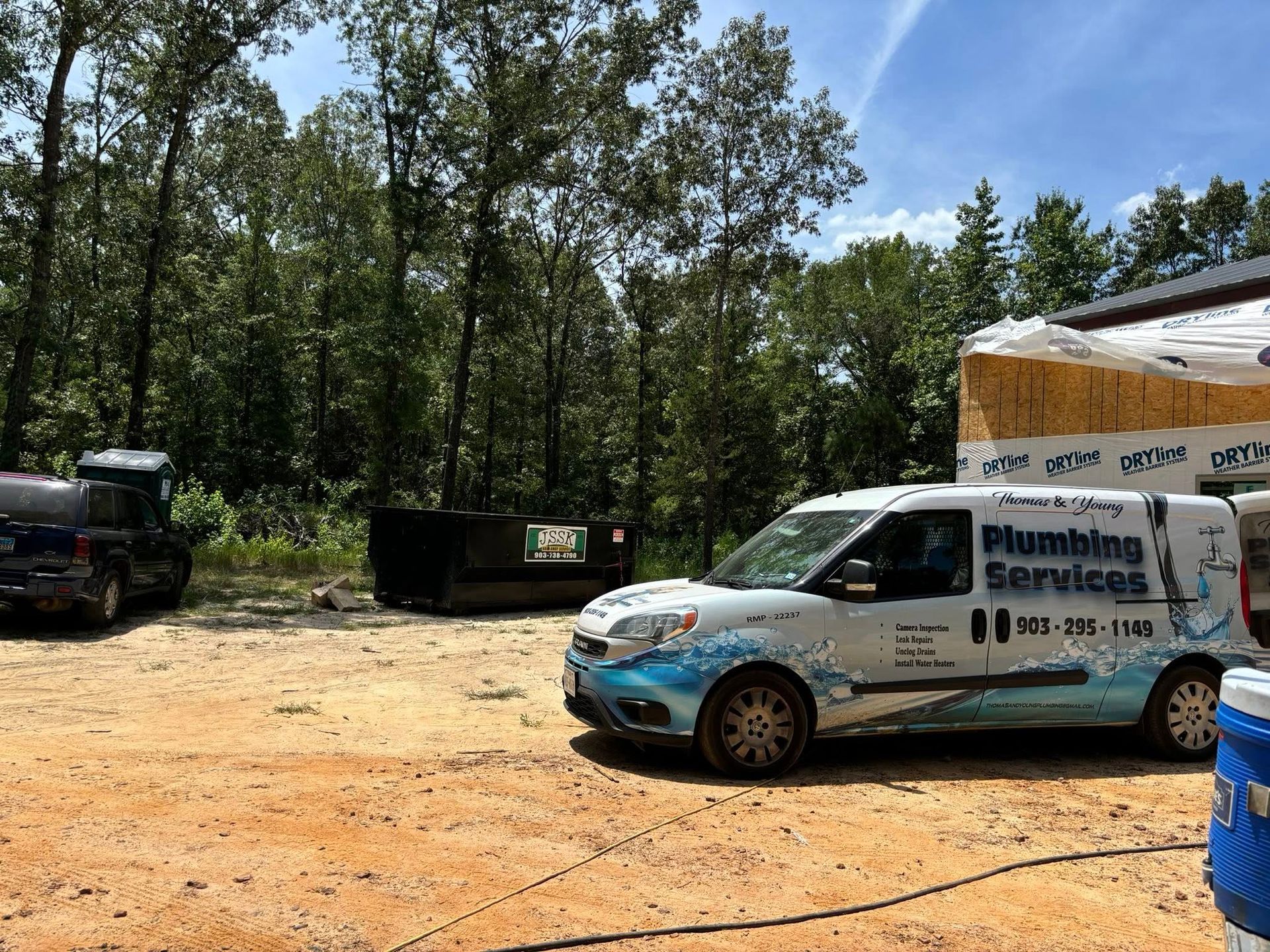 Plumbing services van parked on dirt lot near construction site, trees in background.