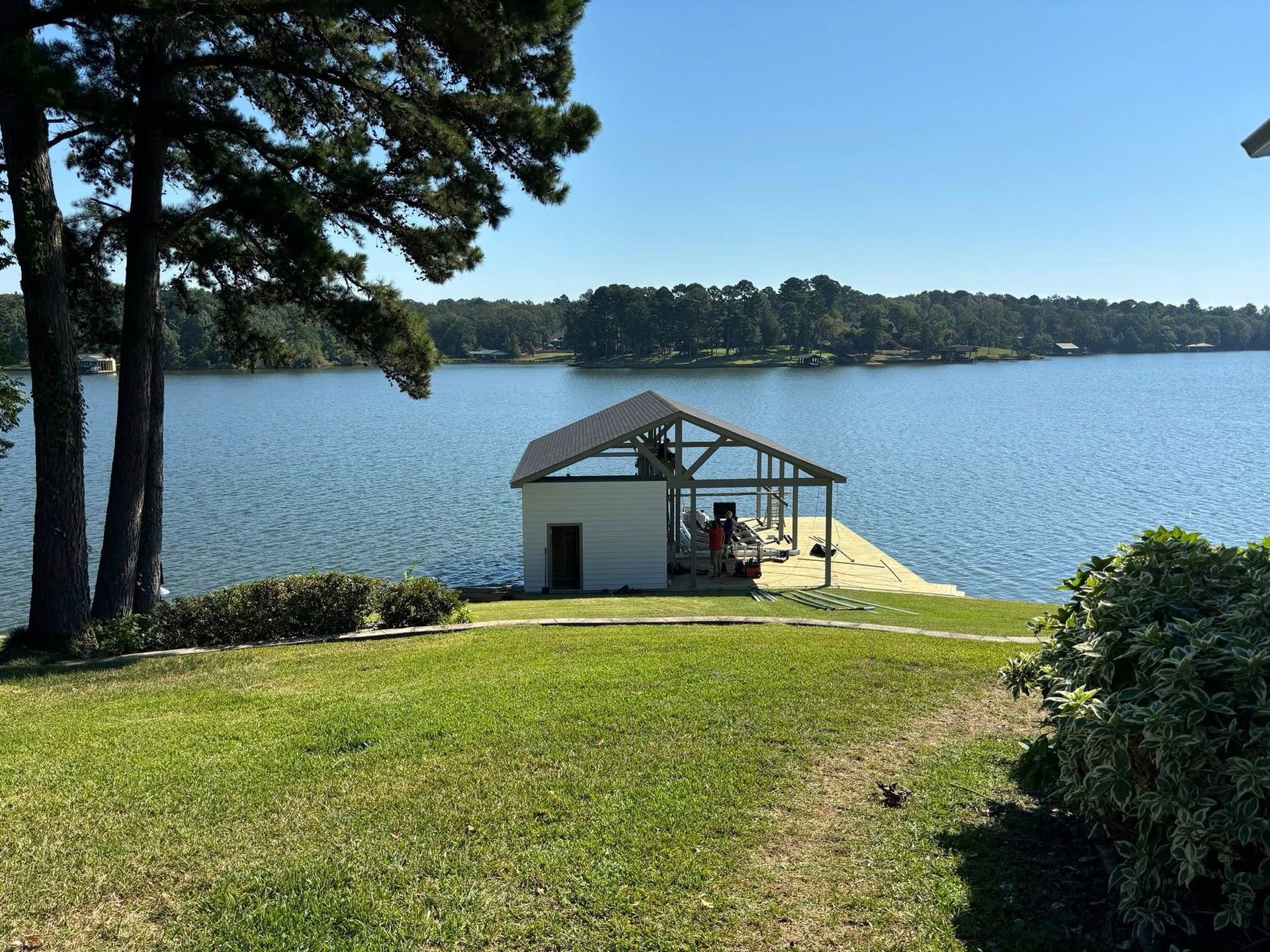 Lakeside boat dock with covered slip, on a sunny day. Green grass in foreground, trees and water in the background.
