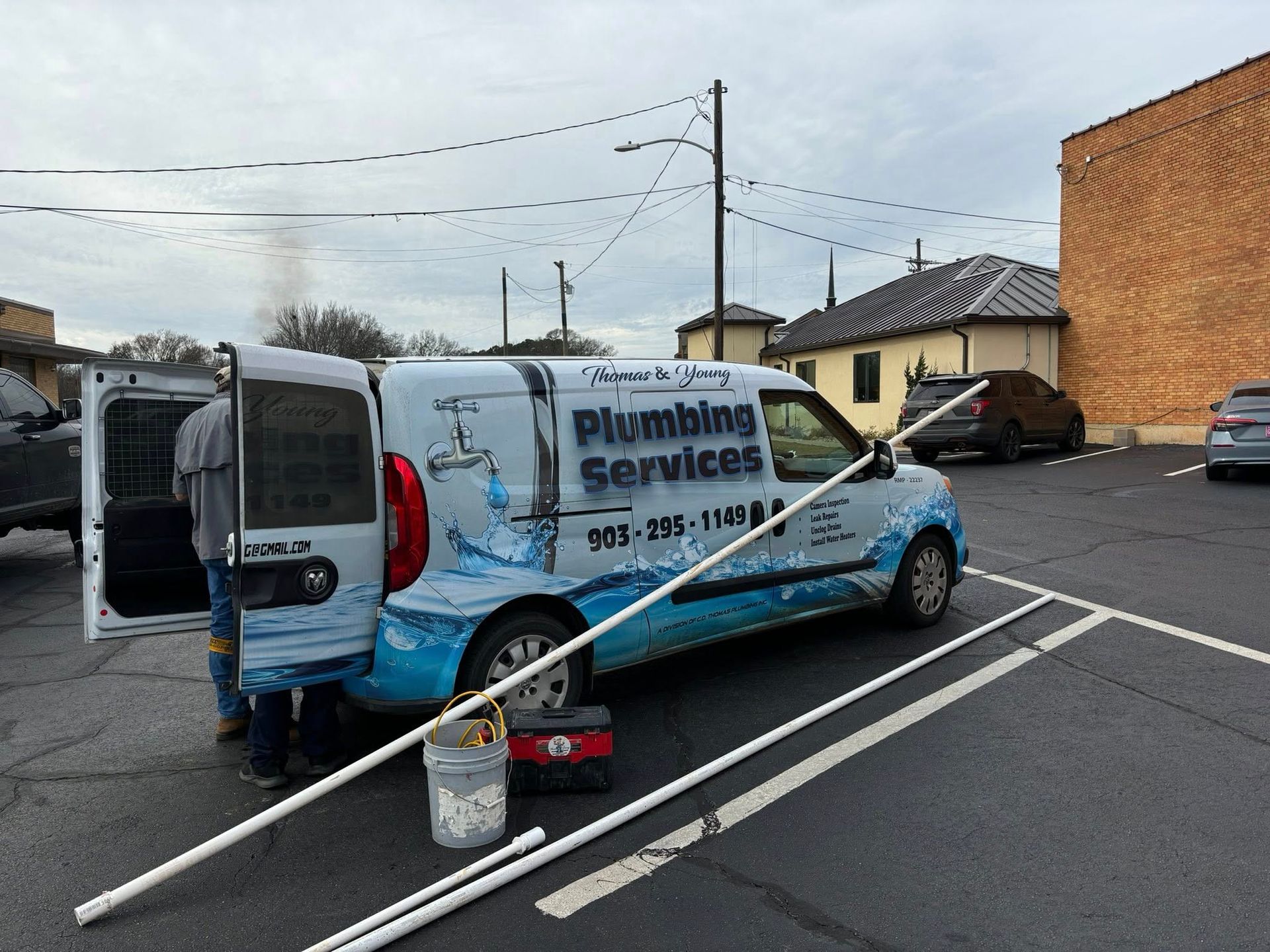 Plumbing services van in parking lot with open door. Worker loads pipes next to a toolbox and bucket. Overcast sky.