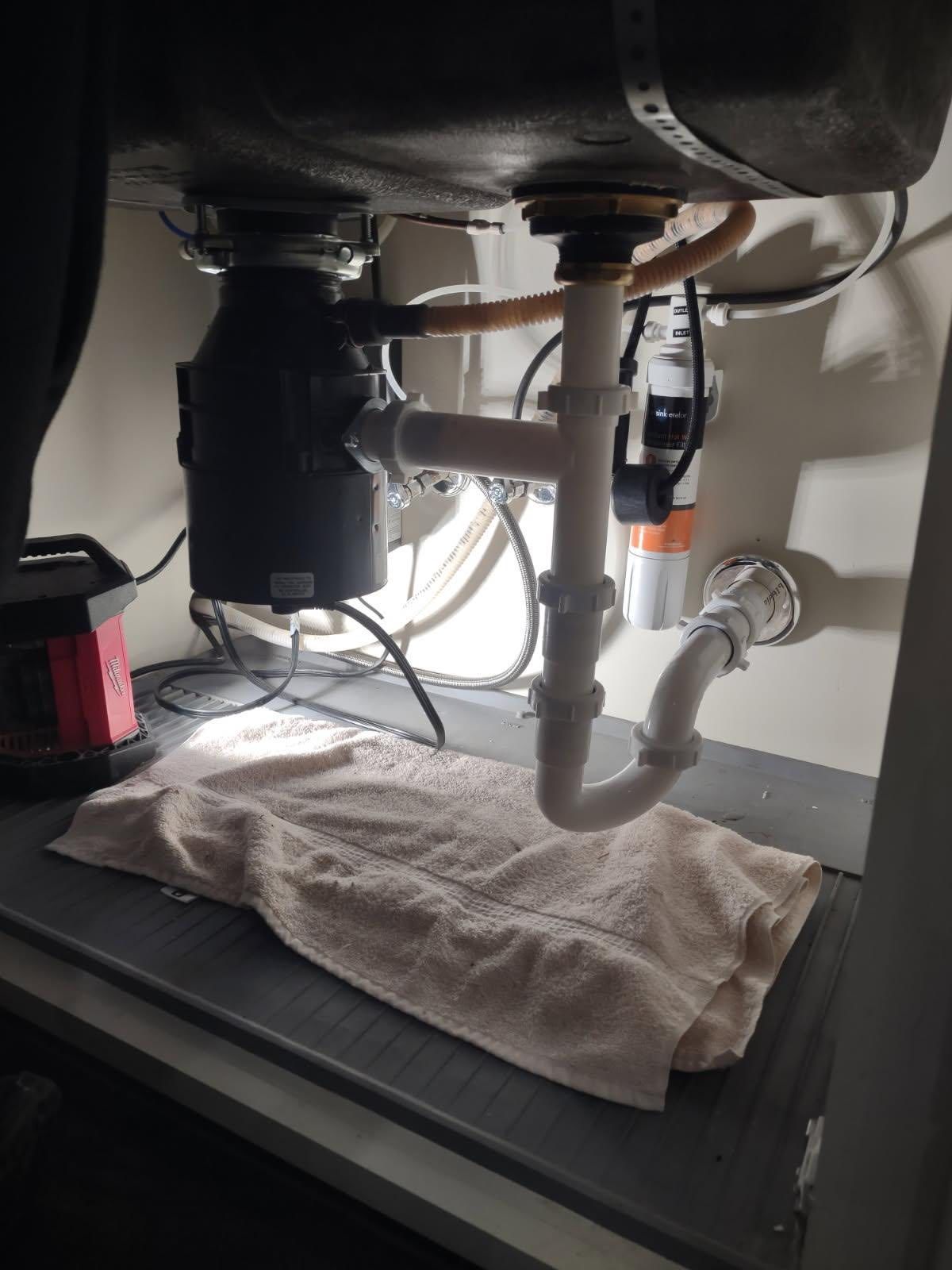Underside of a kitchen sink with a garbage disposal and plumbing pipes; towel on the countertop.