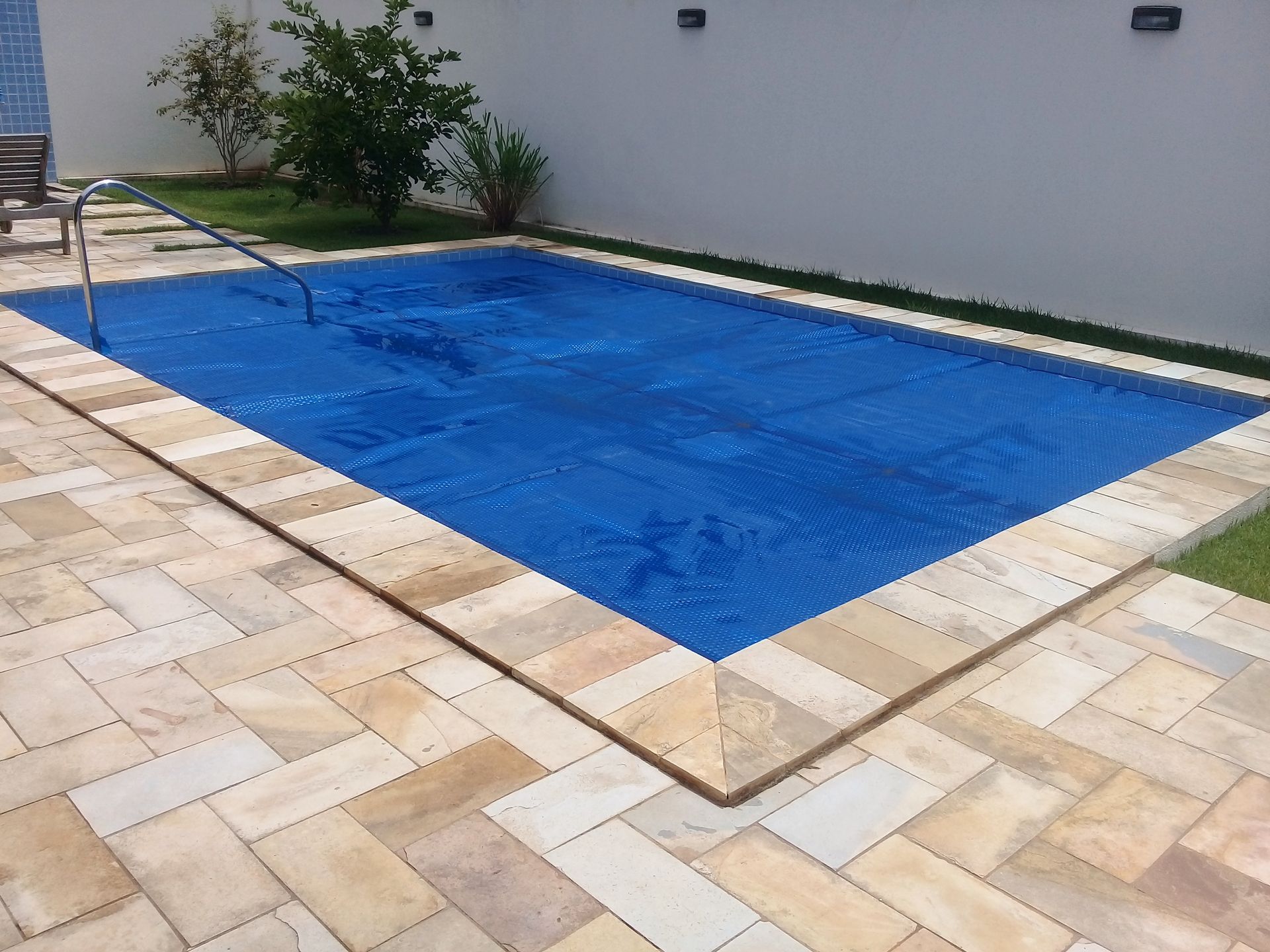 Rectangular pool with blue cover, surrounded by tan stone patio and green grass.