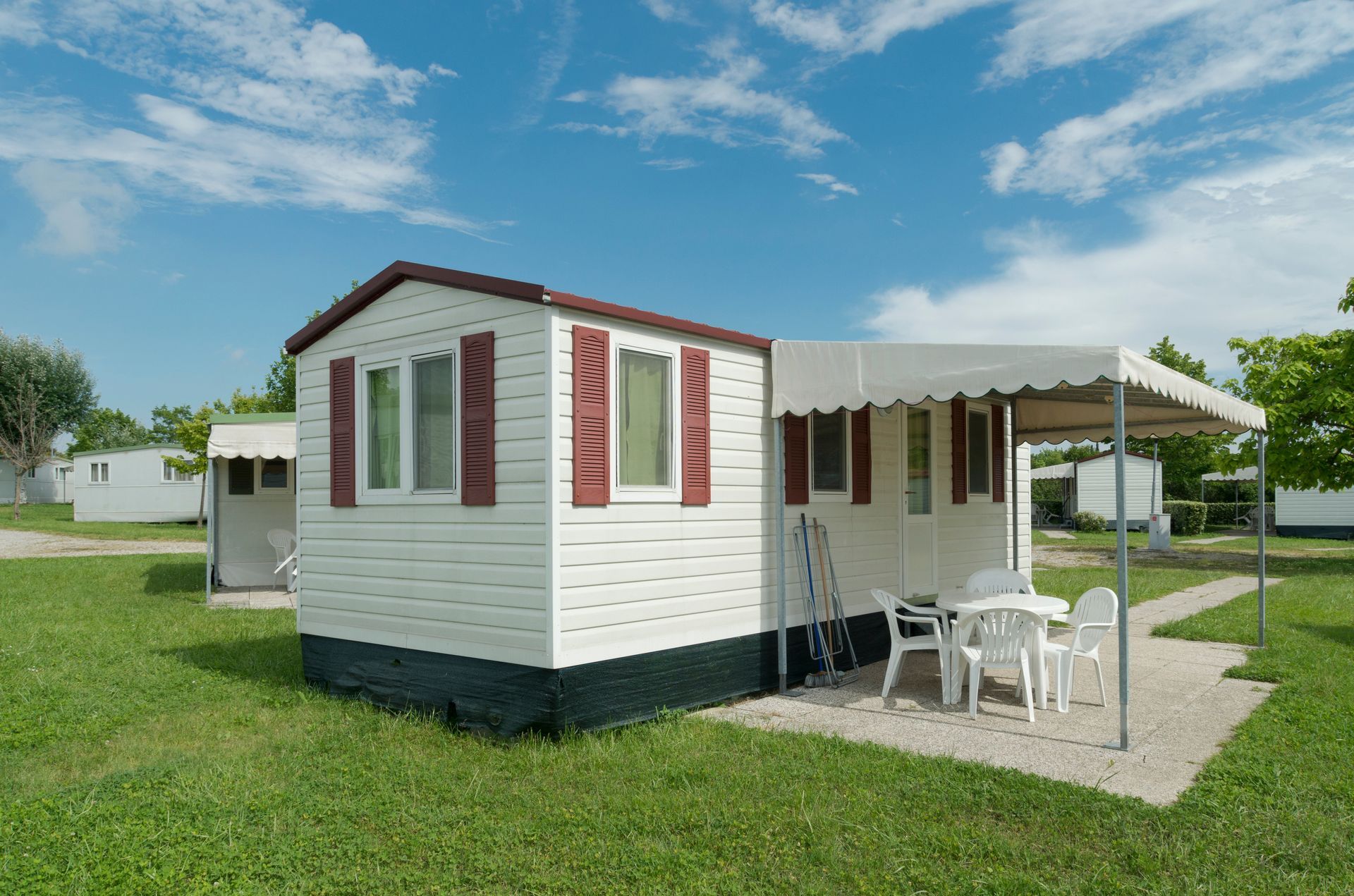 Small white cabin with red-trimmed windows and a patio table under a canopy on a grassy campsite. Small white cabin with red-trimmed windows and a patio table under a canopy on a grassy campsite.