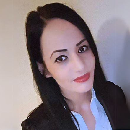 Portrait of a woman with long dark hair, wearing a black jacket and white shirt, smiling slightly against a plain wall