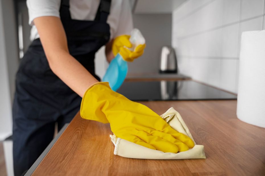 Person in yellow gloves wiping a wooden countertop with a cloth in a kitchen.