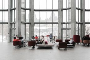 People seated in a bright modern lounge with tall windows and red chairs, overlooking a city view