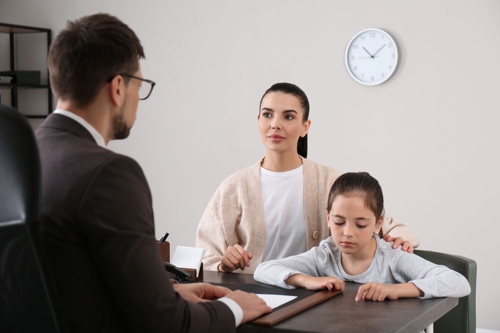Two adults and a child in a meeting room, with the child writing at a desk and a wall clock behind them