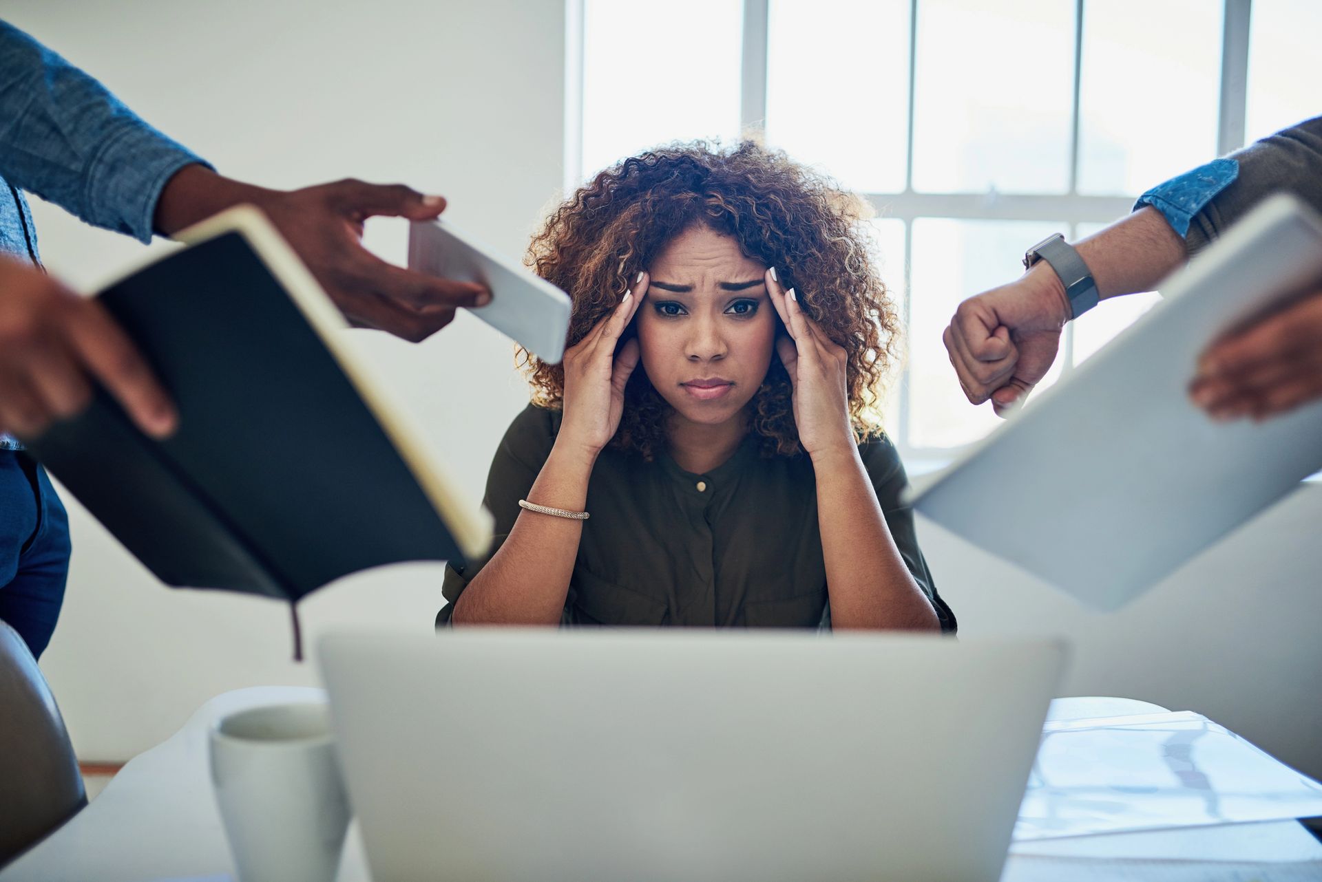 Stressed person at laptop while coworkers point documents at them in a bright office