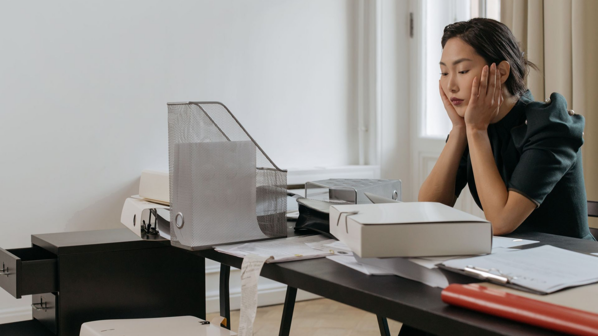Tired woman at a desk with office equipment, resting her head in her hands while looking at paperwork.