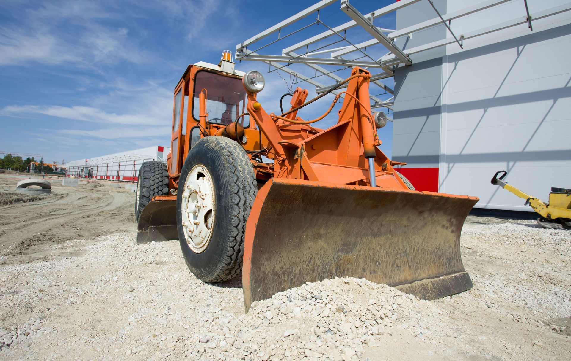 Orange road grader with raised blade on a gravel construction site near a warehouse