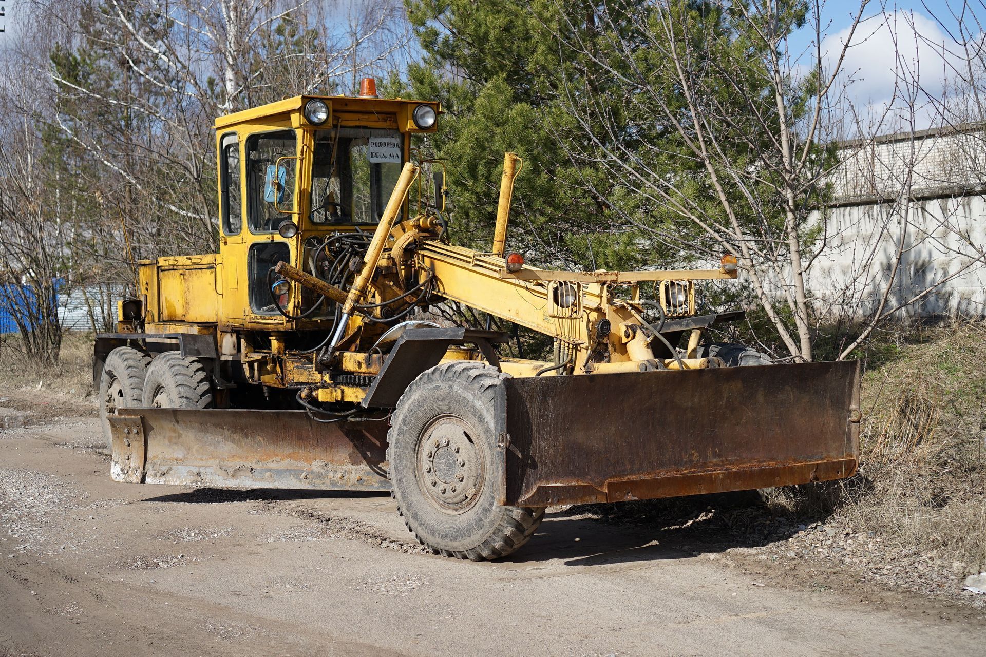 Yellow road grader parked on a dirt road beside trees and a white building