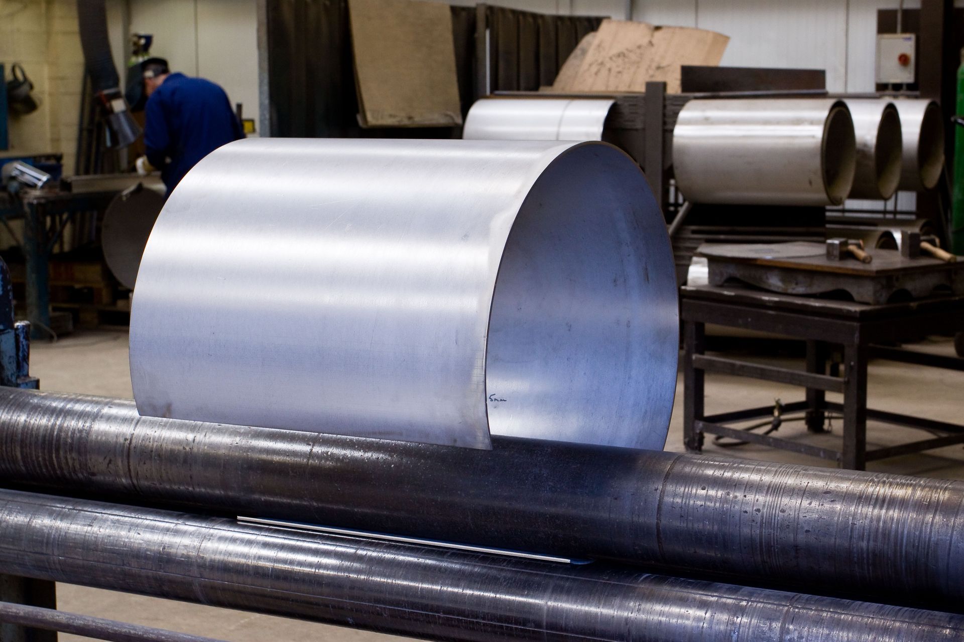 Large curved metal sheet in an industrial workshop, with workers and machinery in the background
