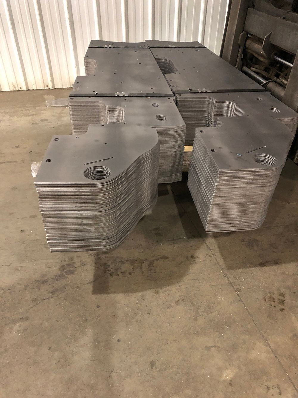 Stacked gray metal plates with holes, arranged on a concrete floor in an industrial area.
