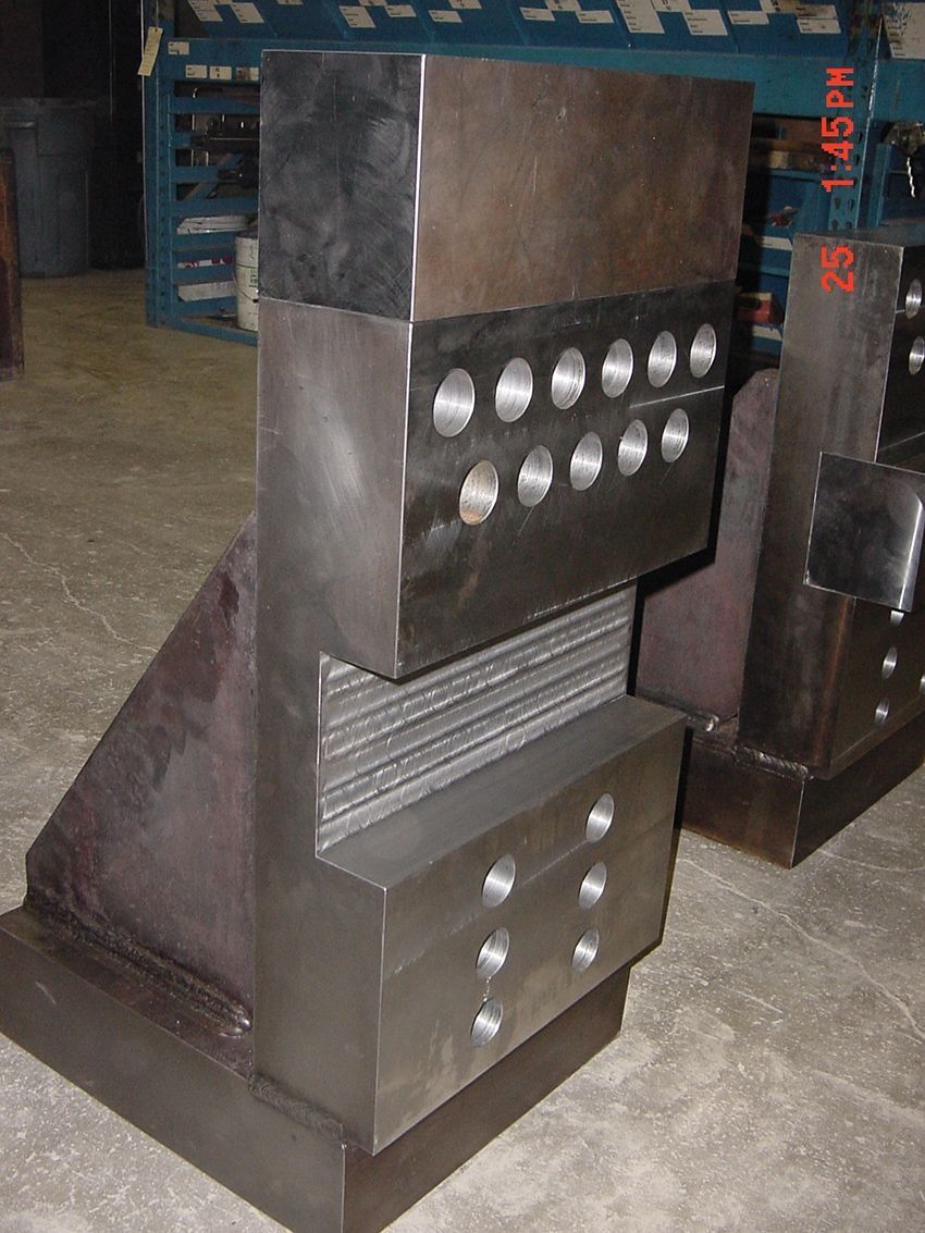Metal industrial block with rows of circular bolts or holes, stacked in a workshop setting