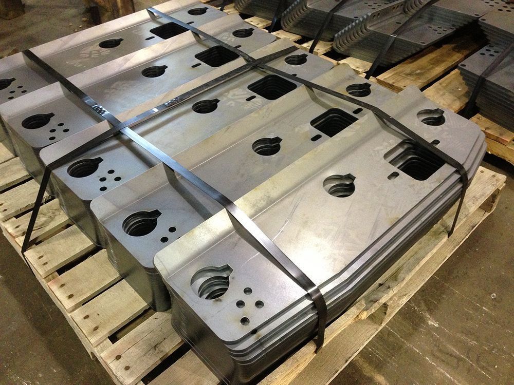 Stack of silver metal plates with cutouts, strapped on a wooden pallet in an industrial warehouse.