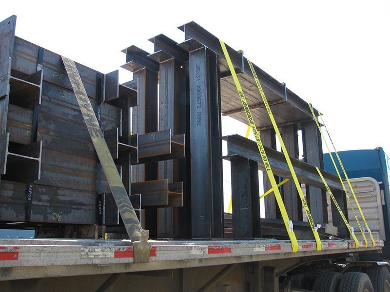Stacked steel beams secured with yellow straps on a flatbed trailer.