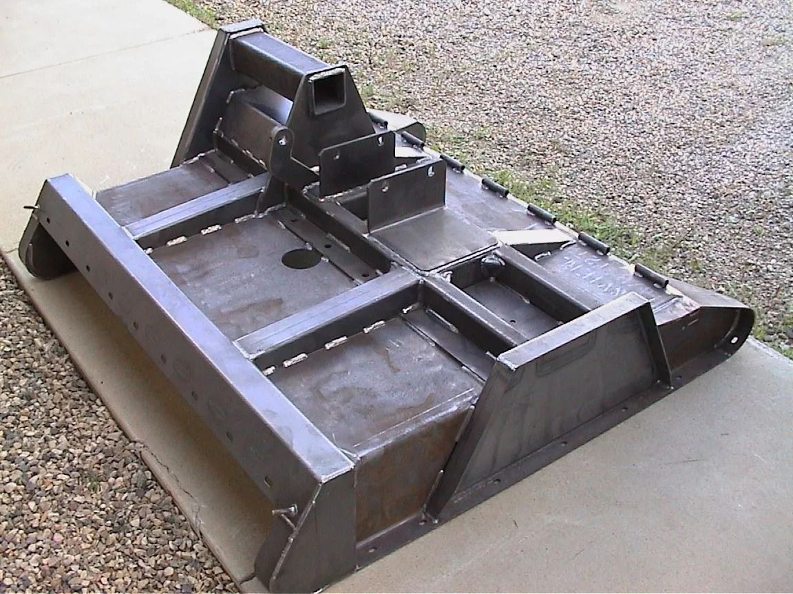Black metal mower deck attachment resting on gravel, with raised central housing and side guard panels