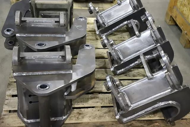 Stacked silver metal brackets and machined parts on wooden pallets in a workshop or warehouse.