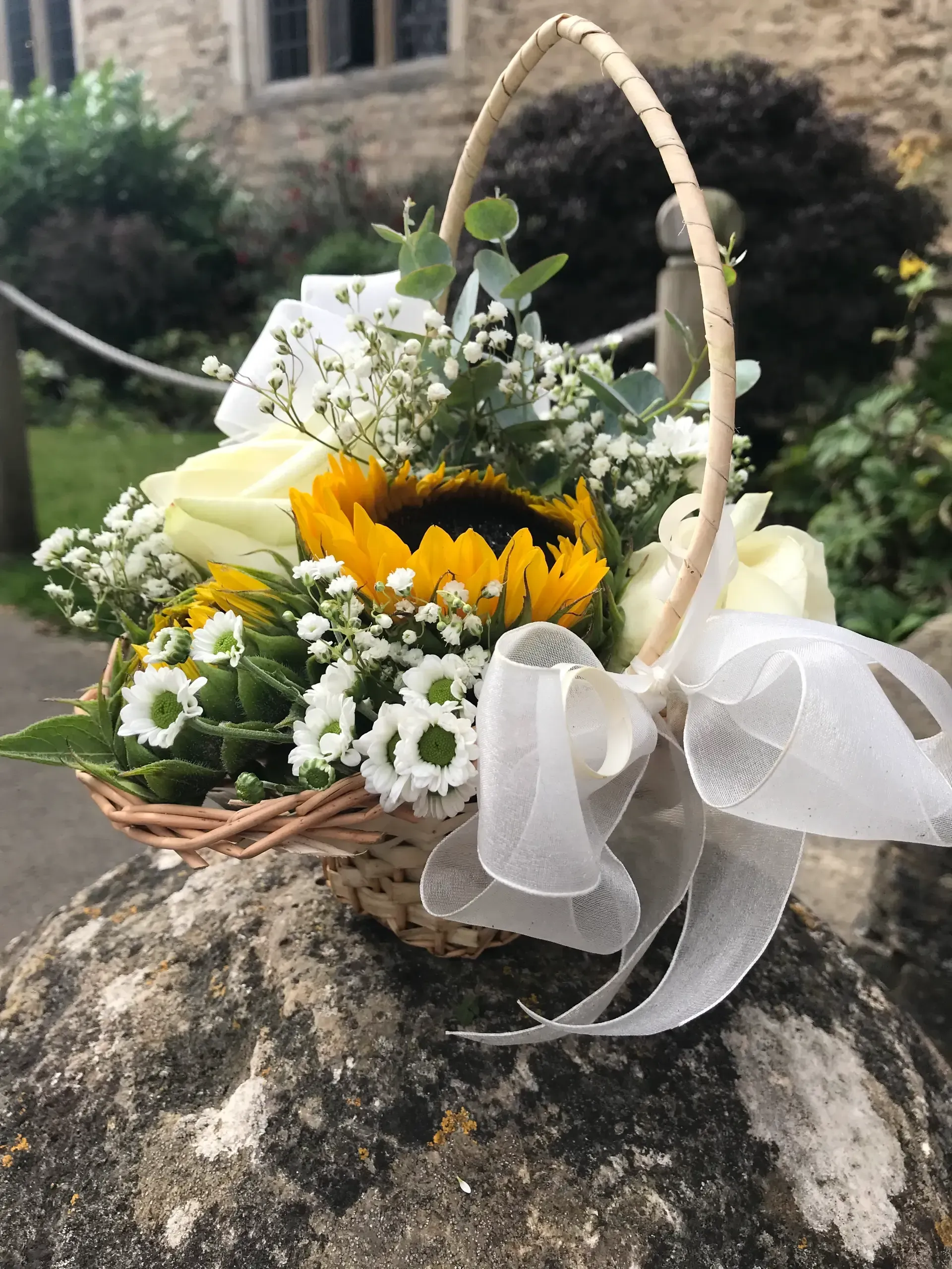 Small basket of white roses and yellow sunflower with a white ribbon on a stone outdoors