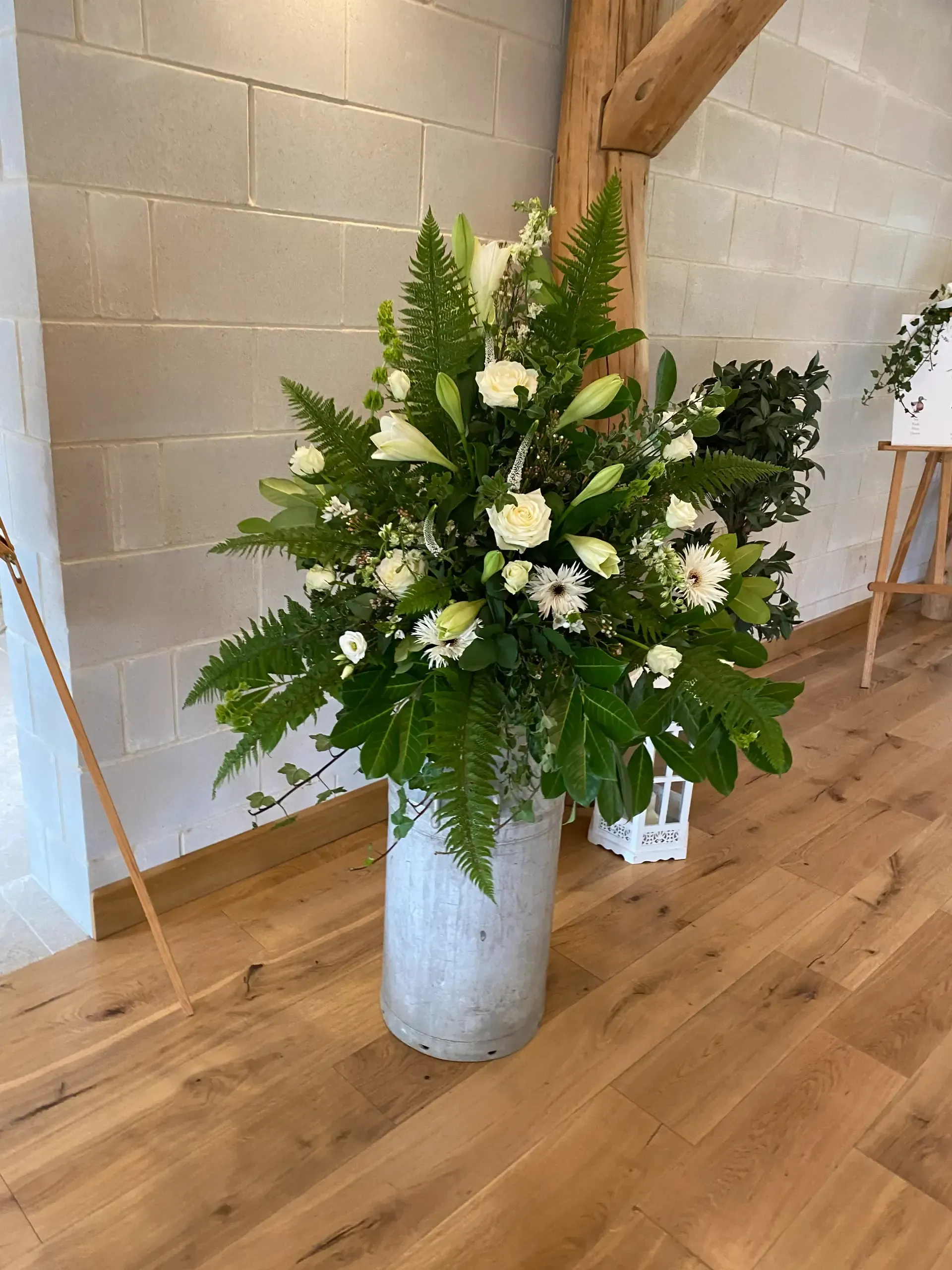 Large white and green floral arrangement in a silver vase on a wooden floor against a light brick wall