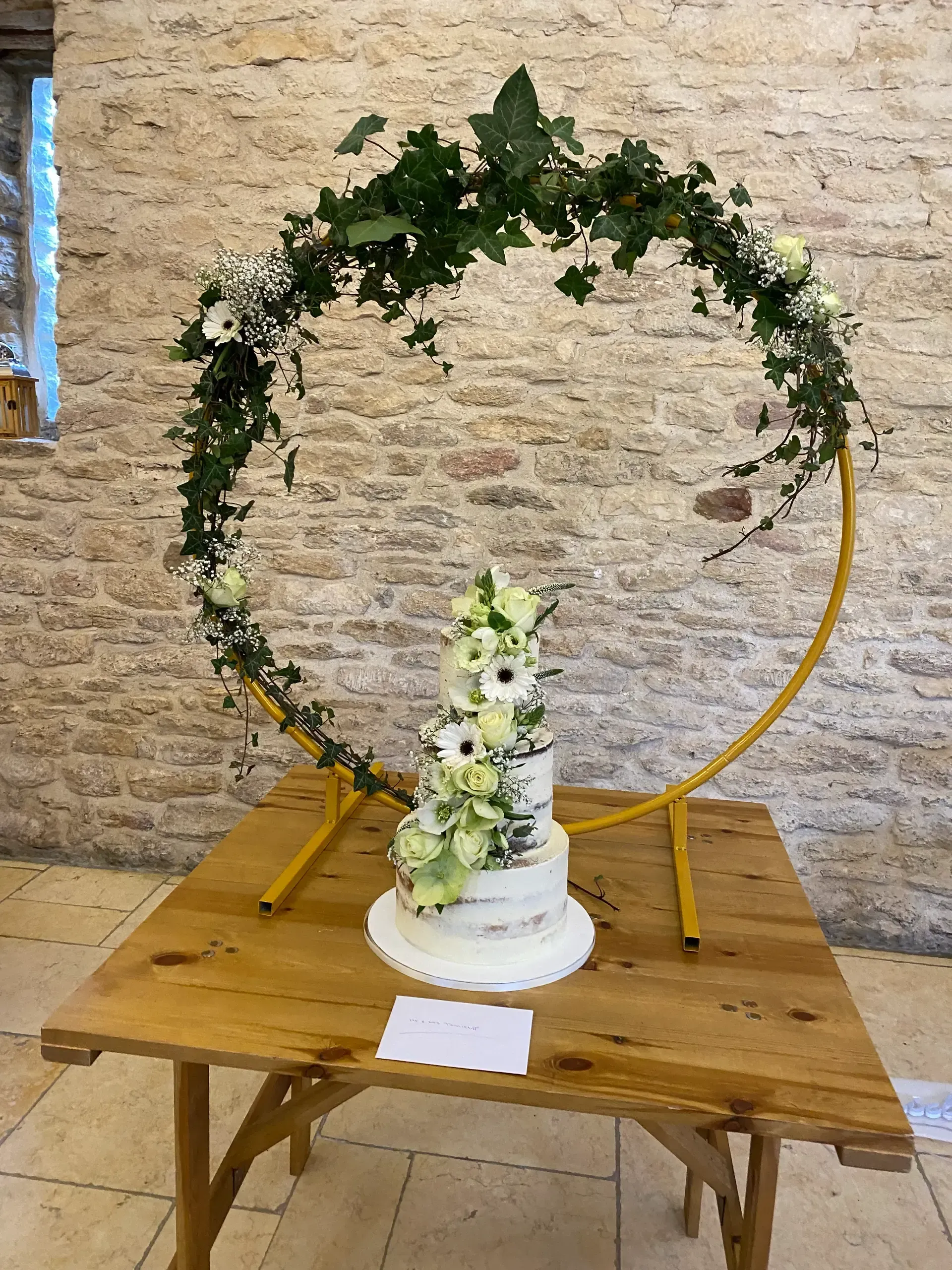 White flower arrangement on a wooden table beneath a large circular greenery arch against a stone wall.