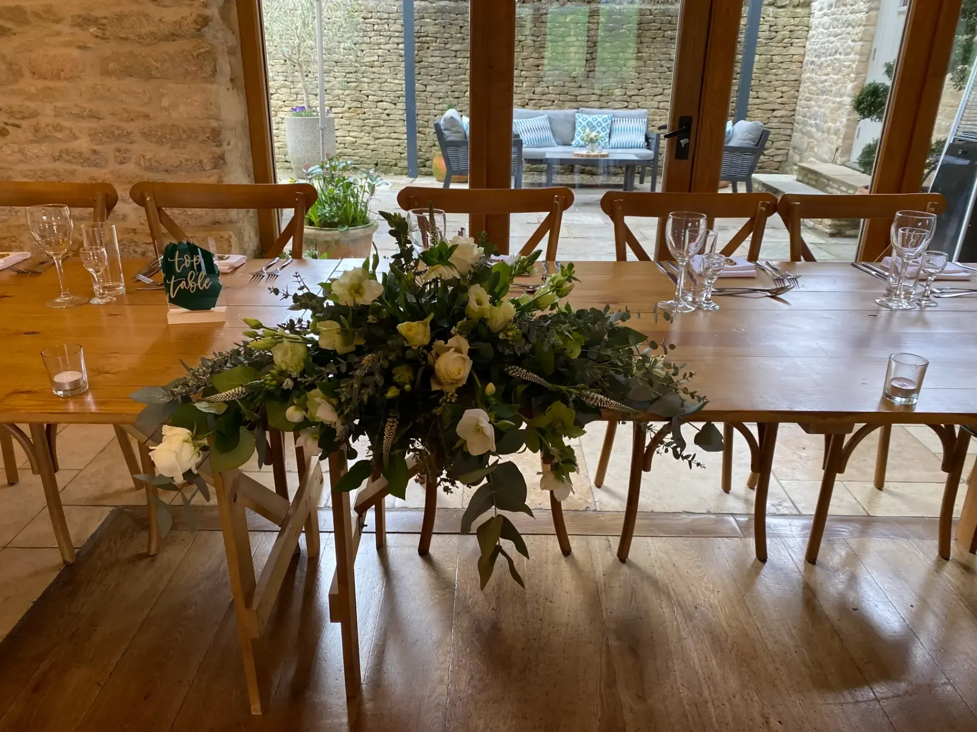 Elegant banquet table with white chairs, floral centerpiece, and glassware in a rustic room with large windows