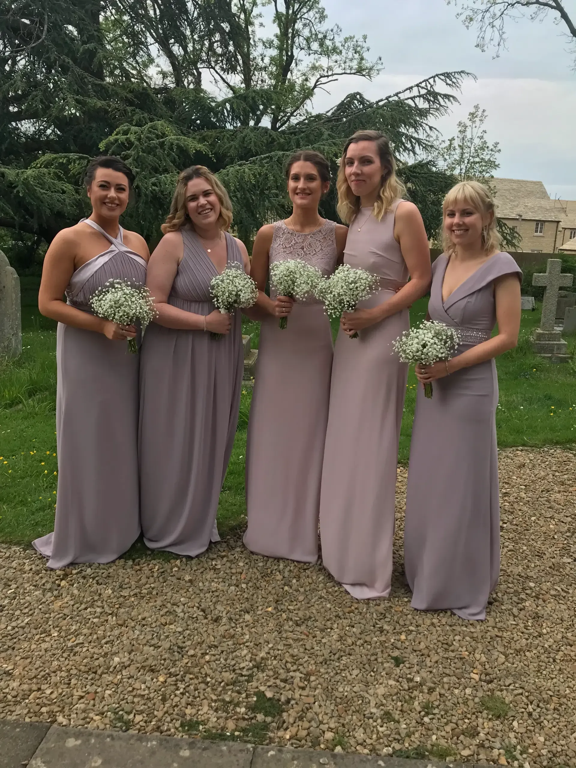 Five women in lavender dresses holding bouquets stand outdoors on gravel under cloudy skies.