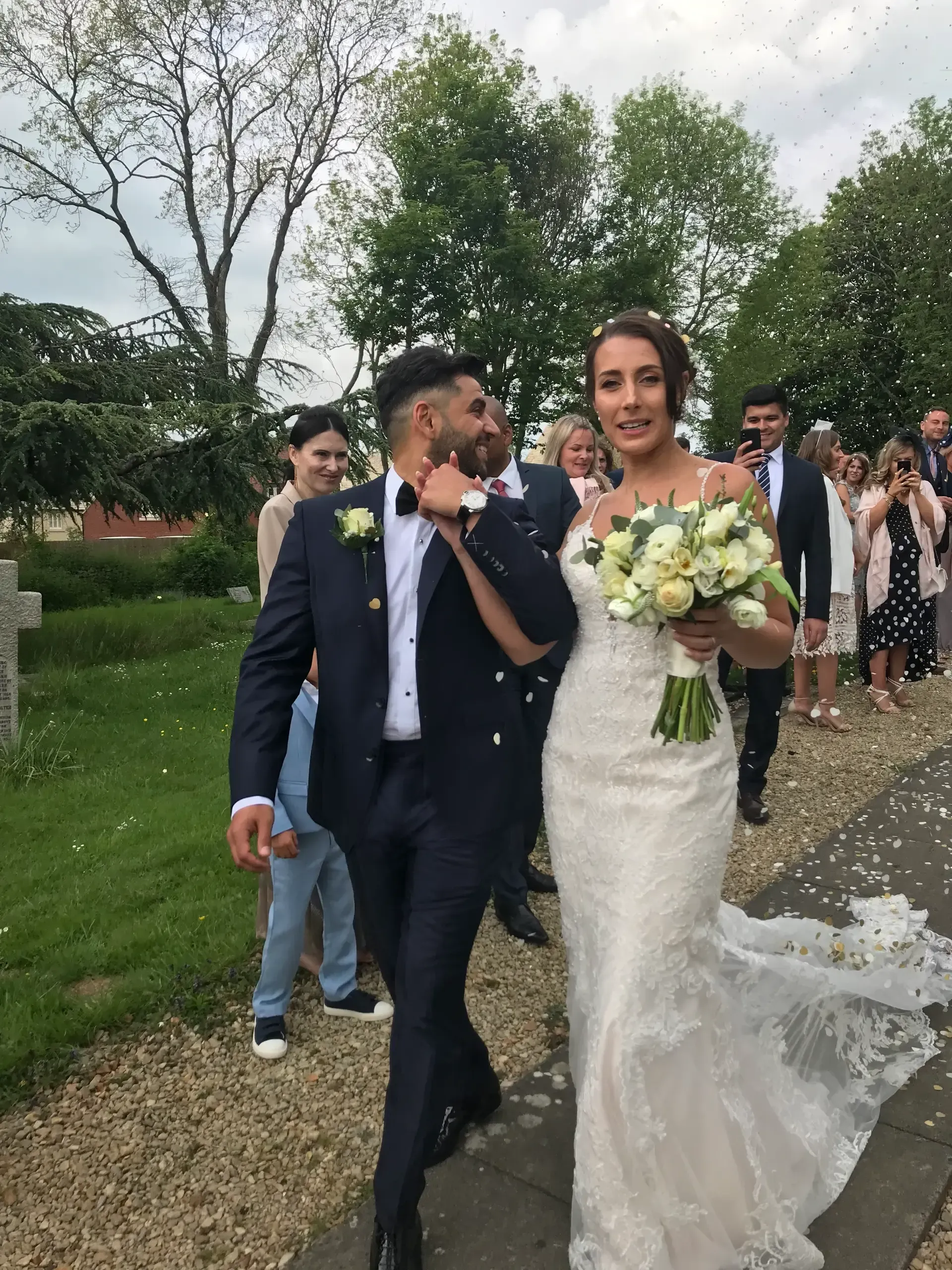 Bride in white dress and groom in navy suit walking outdoors, smiling and holding a bouquet.