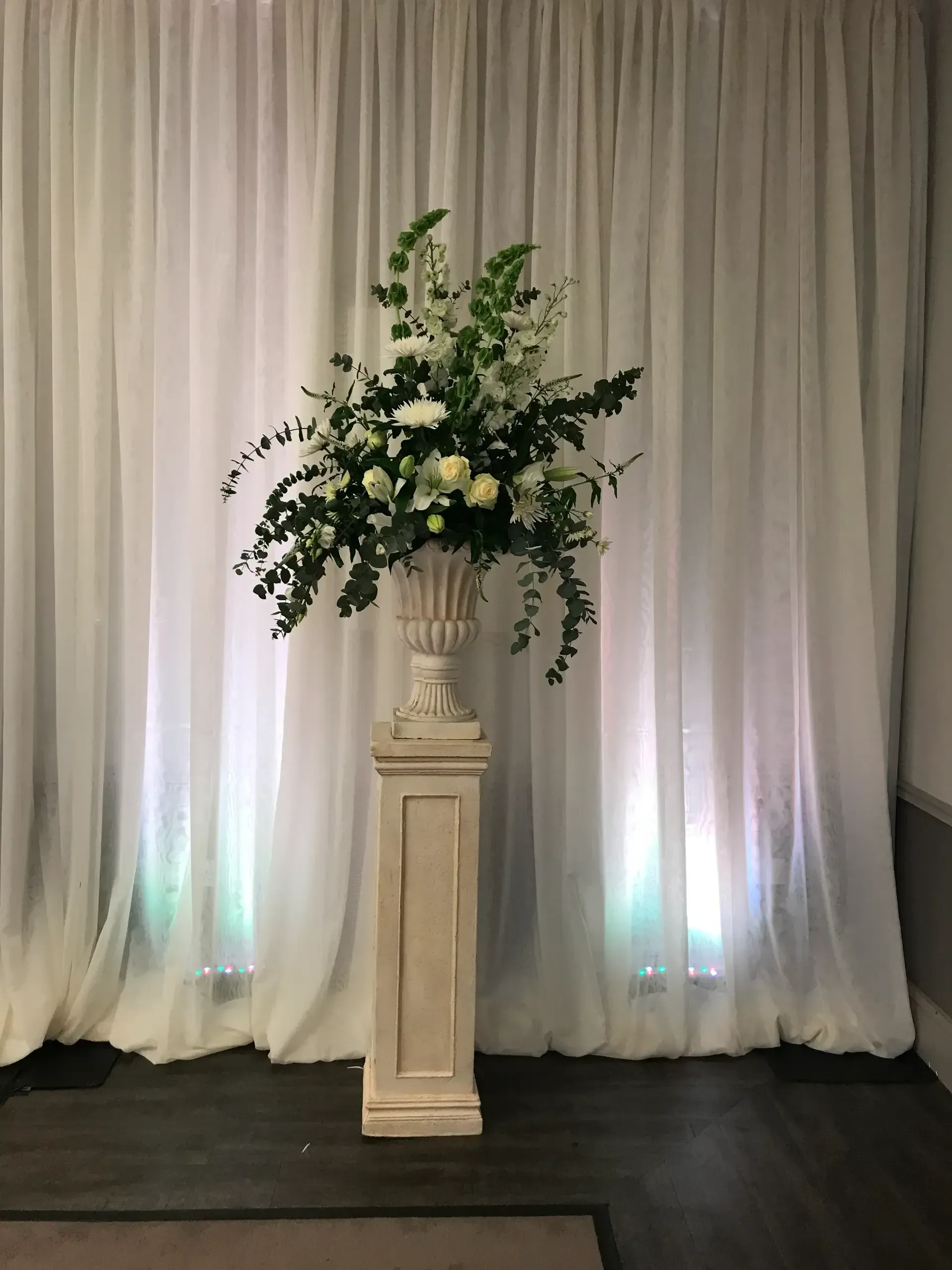 Floral arrangement on a white pedestal before draped white curtains with soft colored uplighting