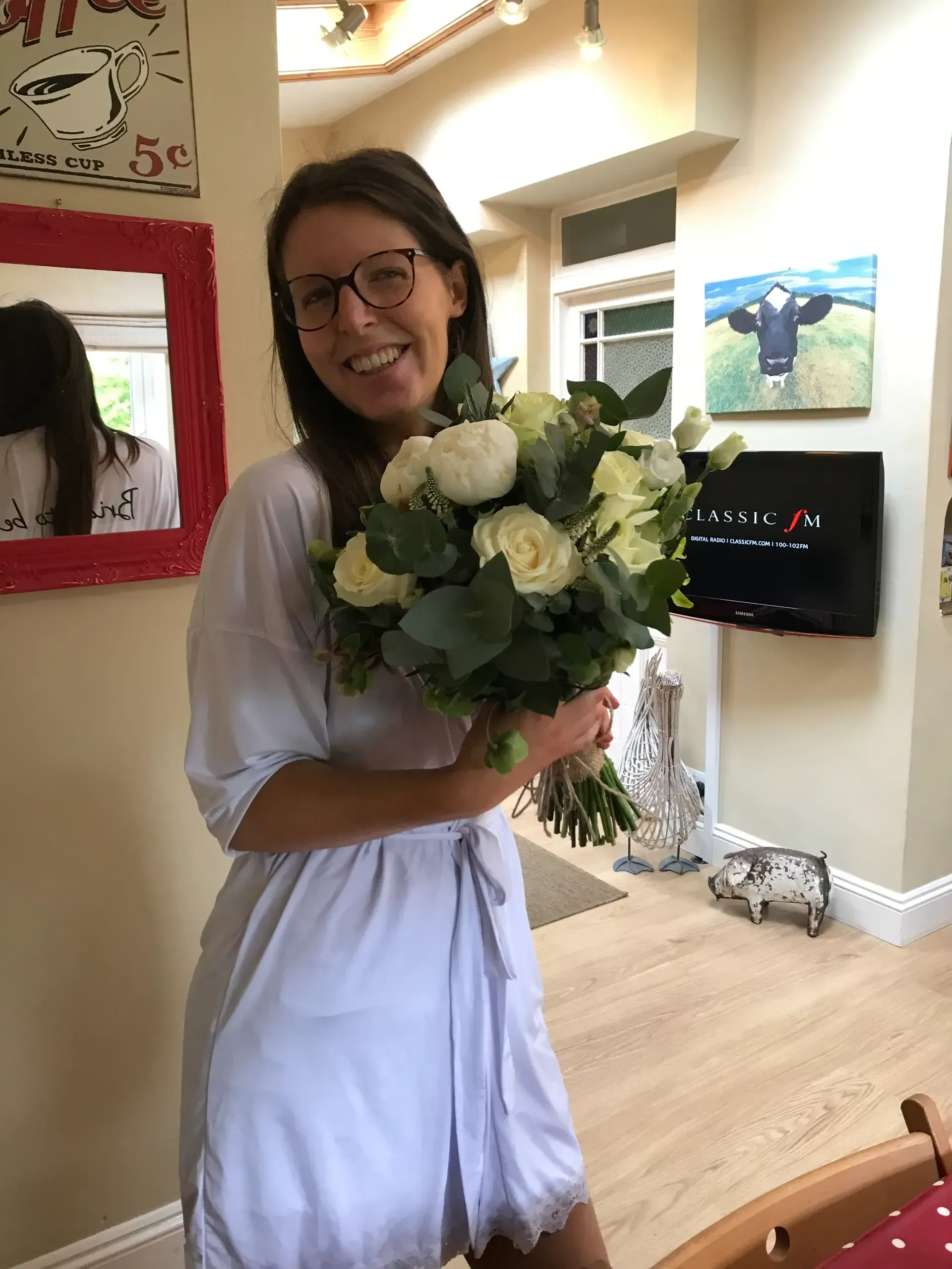 Smiling woman in a white dress holding a bouquet of white flowers in a bright room.