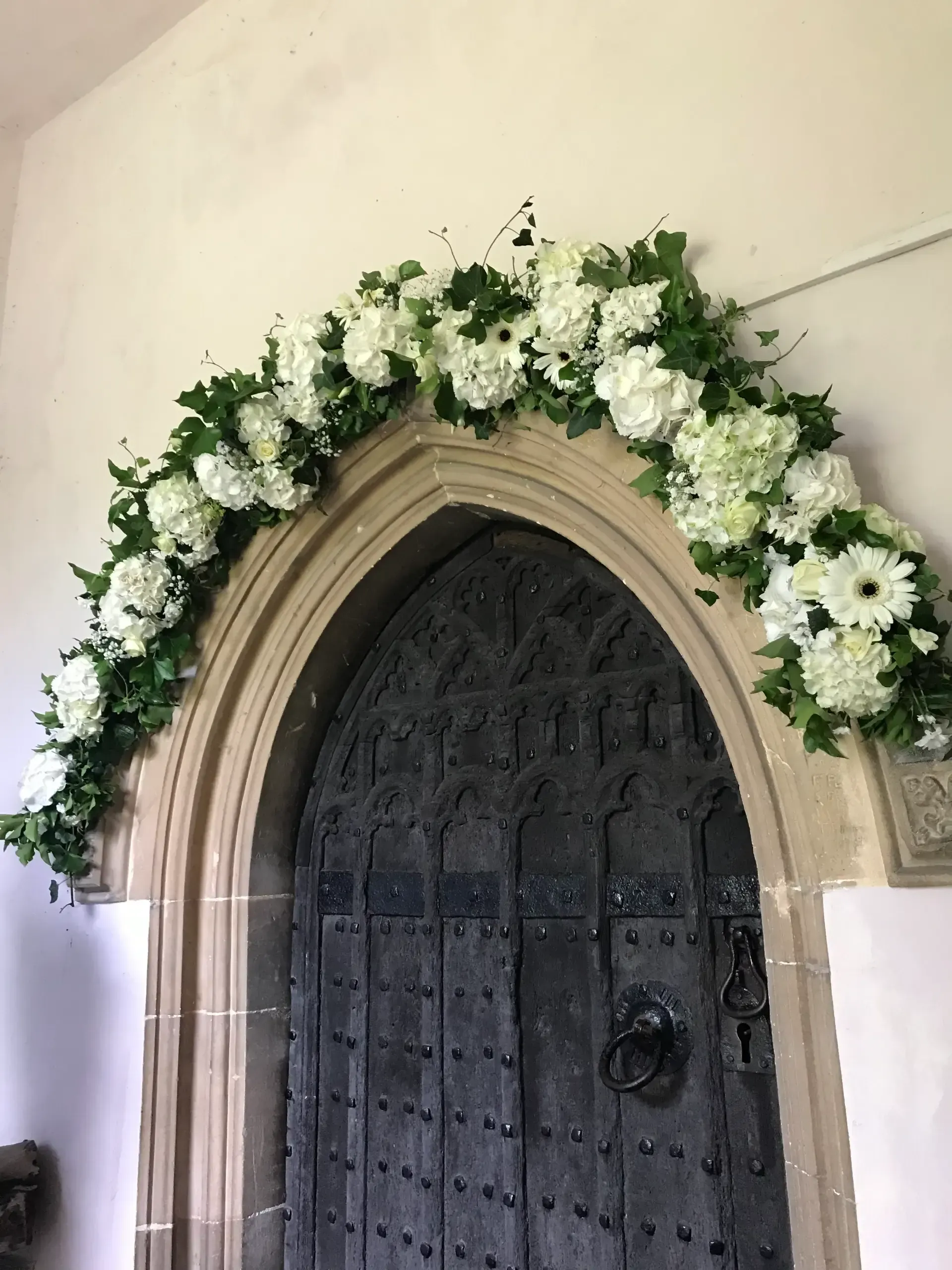 White flower garland arch over a dark wooden church door in a stone doorway
