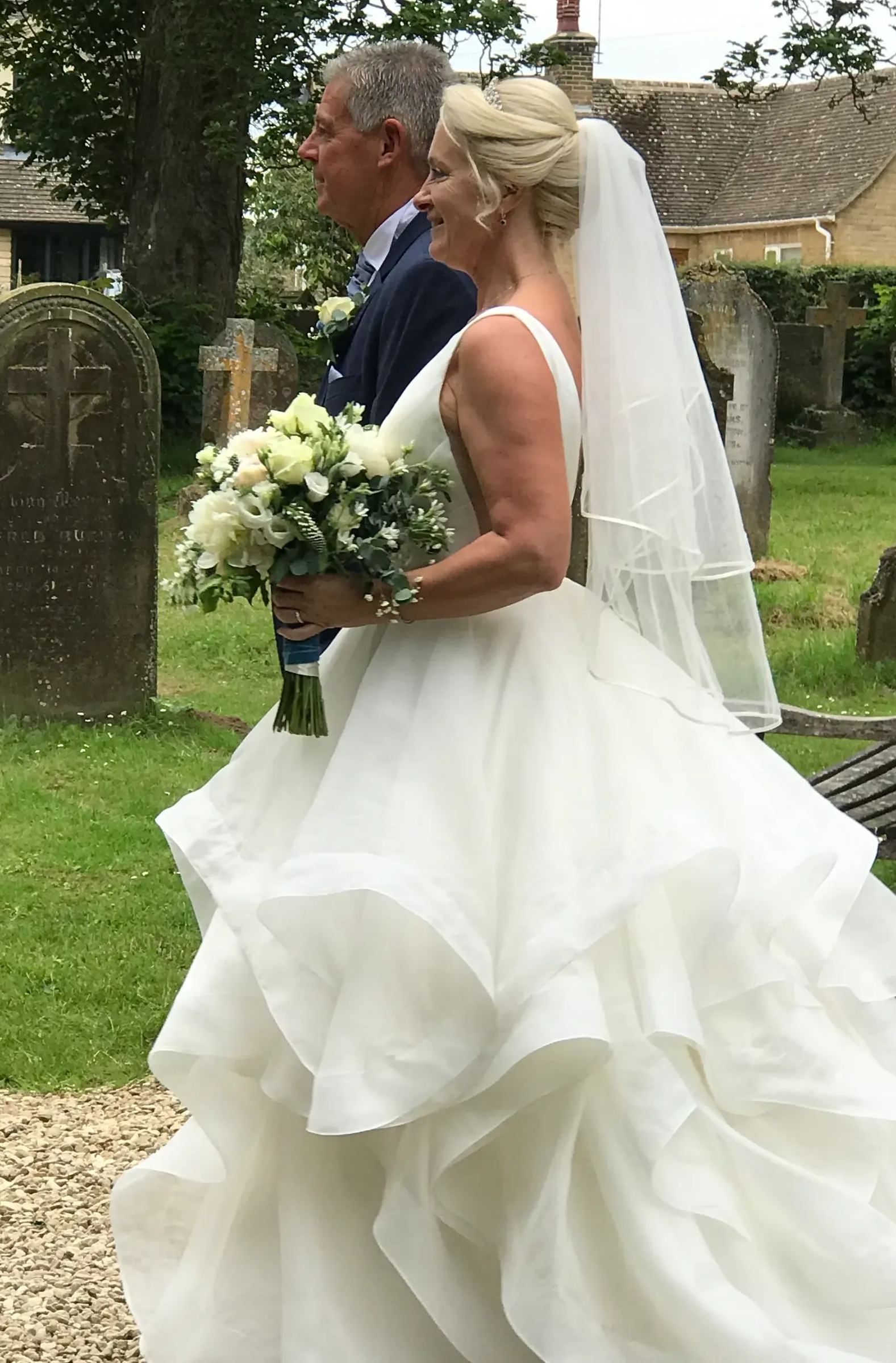 Bride in a white gown and veil walks beside a man outdoors, holding a bouquet.