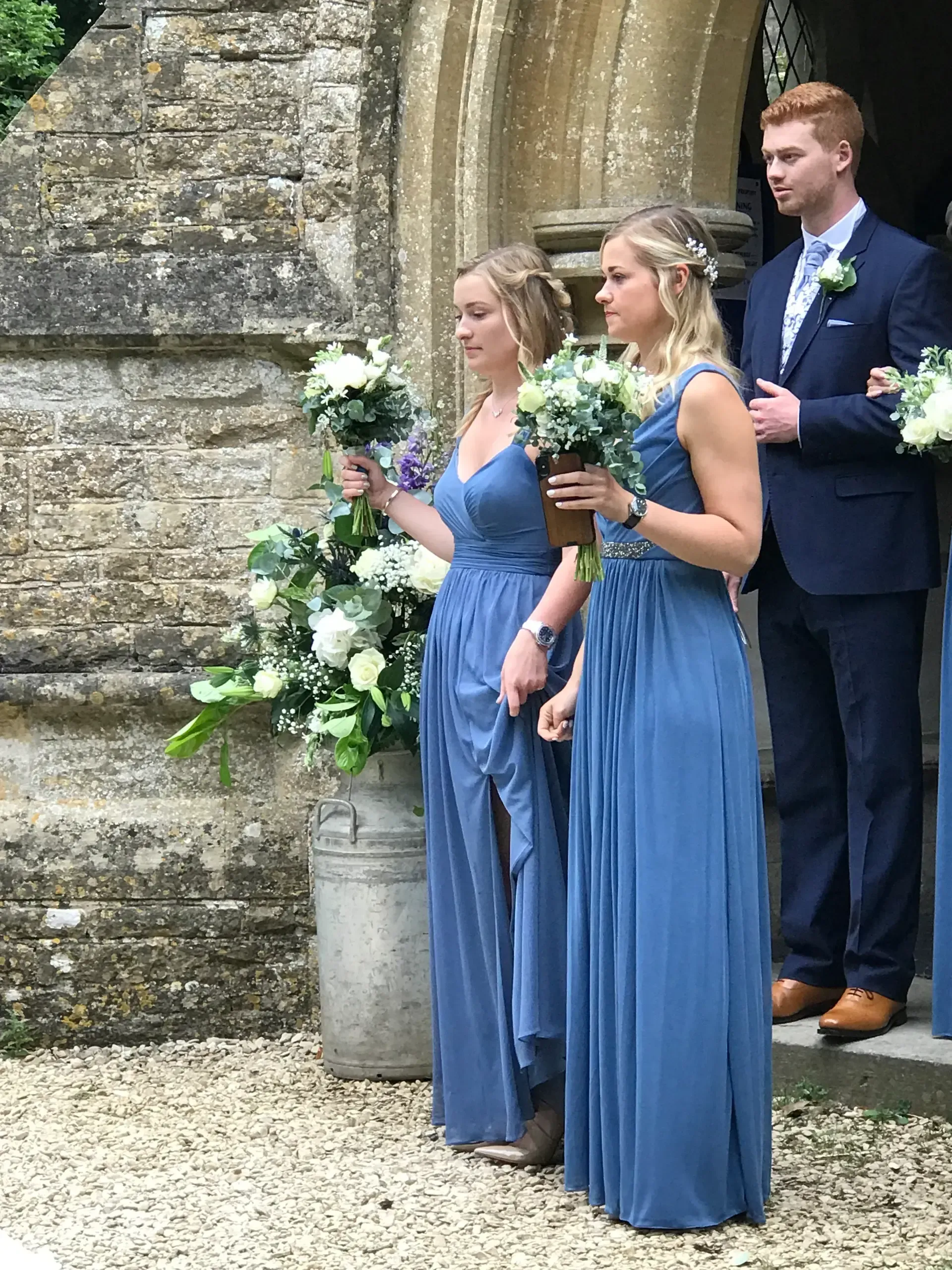 Three people in blue formal attire stand outdoors, two holding bouquets beside a stone wall.