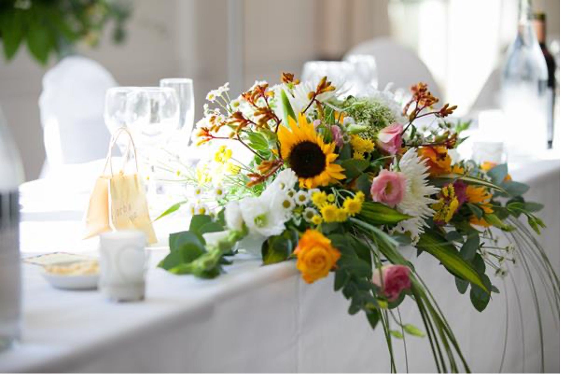 White banquet table with a bright sunflower and mixed flower centerpiece, set with glassware and candles