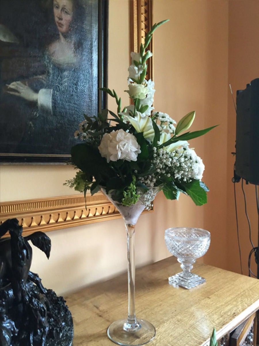 Tall floral arrangement with white flowers and greenery on a wooden table beside a crystal bowl and portrait painting