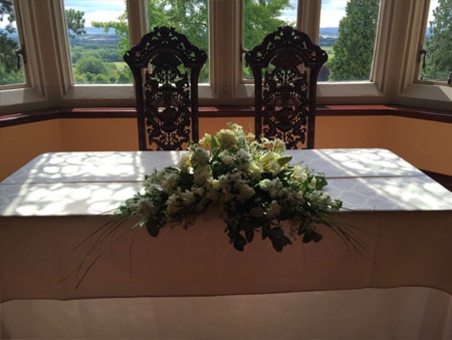Sunlit table with white cloth and floral centerpiece in front of two ornate chairs and windows
