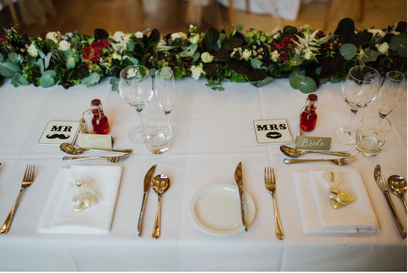 Elegant wedding reception table setting with white plates, gold silverware, and floral greenery centerpiece