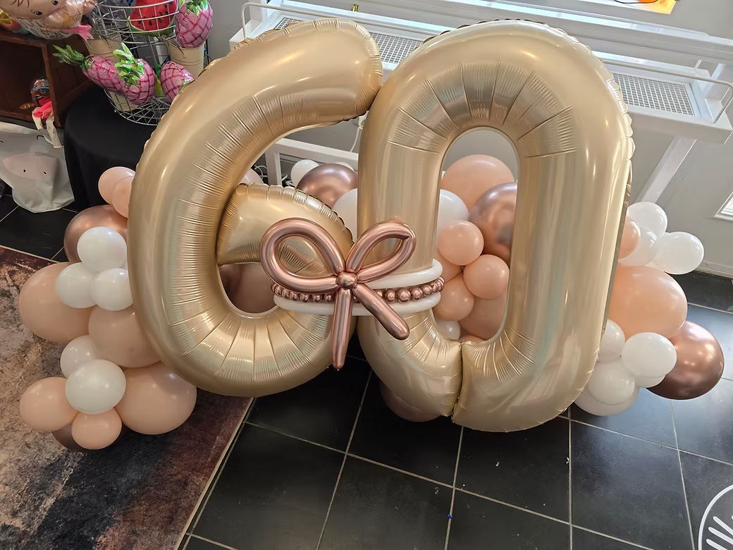 Gold balloon monogram with pink ribbon, surrounded by peach and white balloons on a tiled floor
