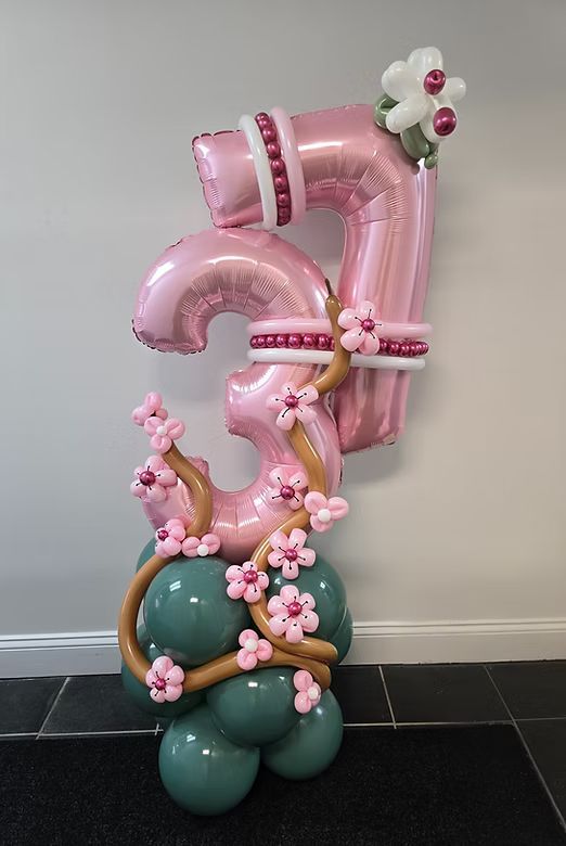 Pink balloon number 3 with floral accents on a green balloon base against a gray wall