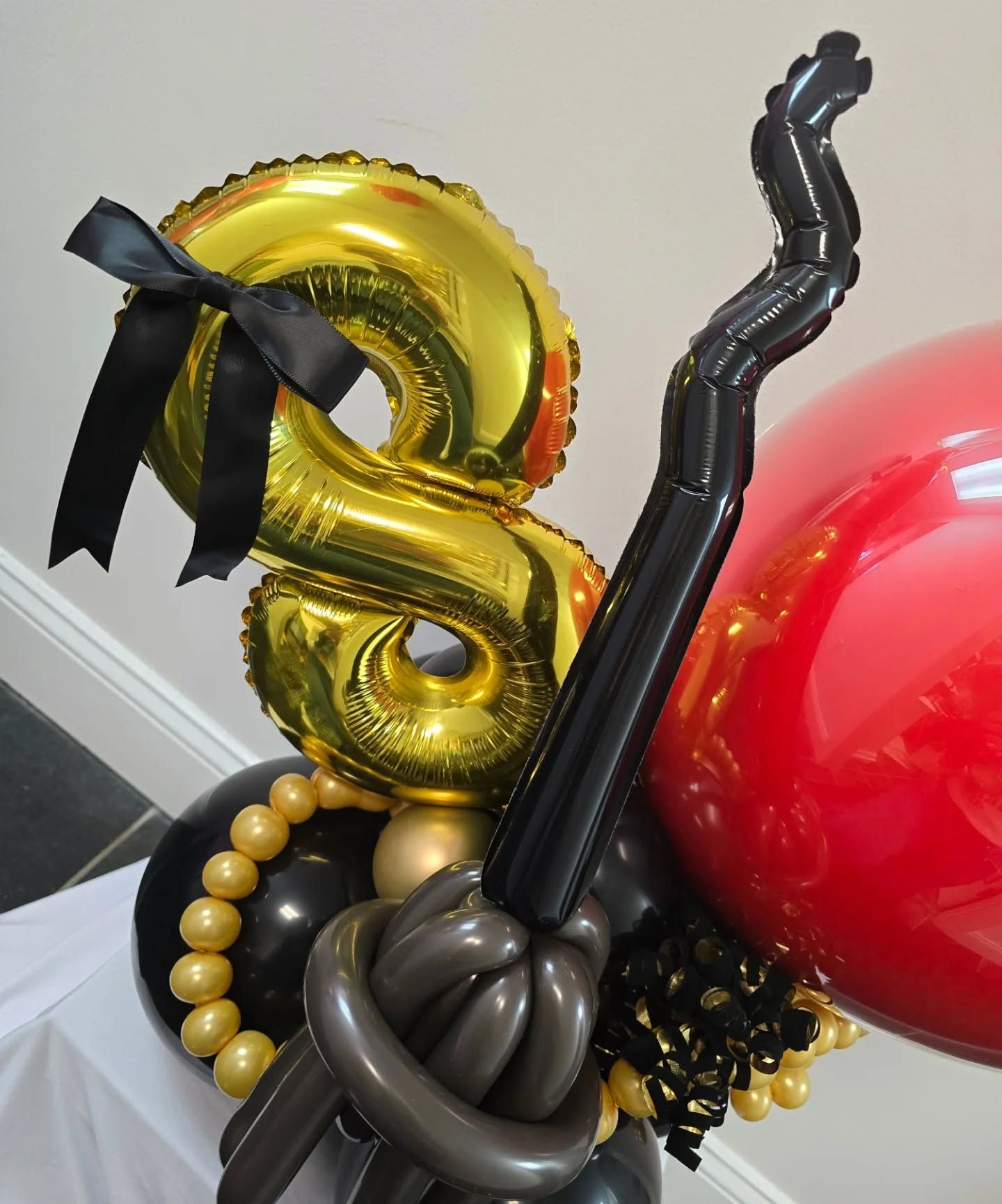Black and gold balloon arrangement with a shiny gold bow and red balloons in the background