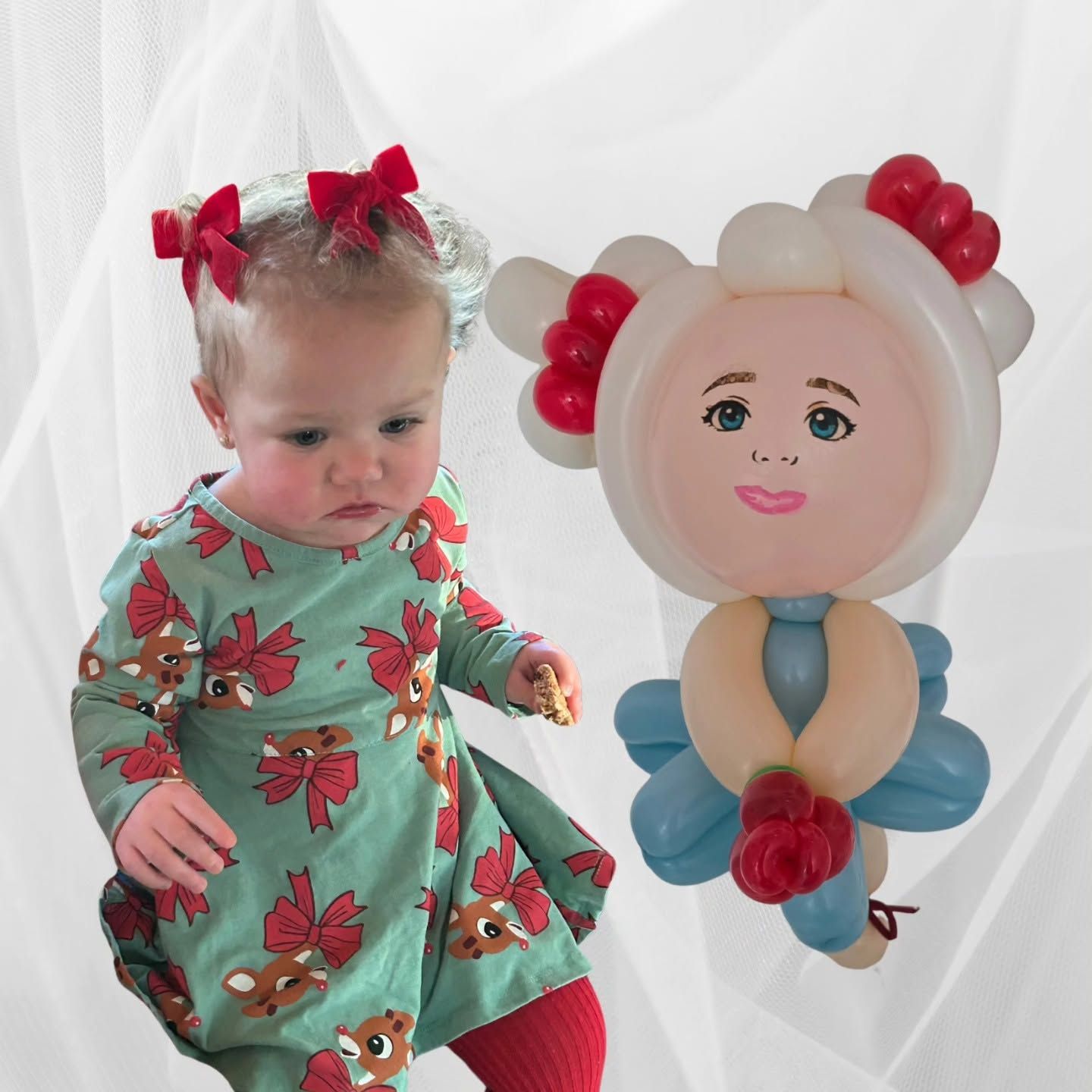 Baby in green holiday dress beside a balloon flower with a smiling face and red petals