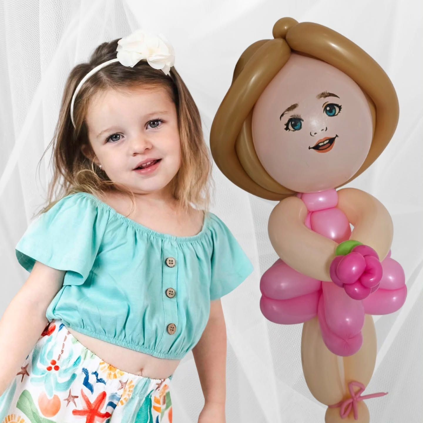 Young child smiling beside a pink-and-beige balloon doll against a white backdrop